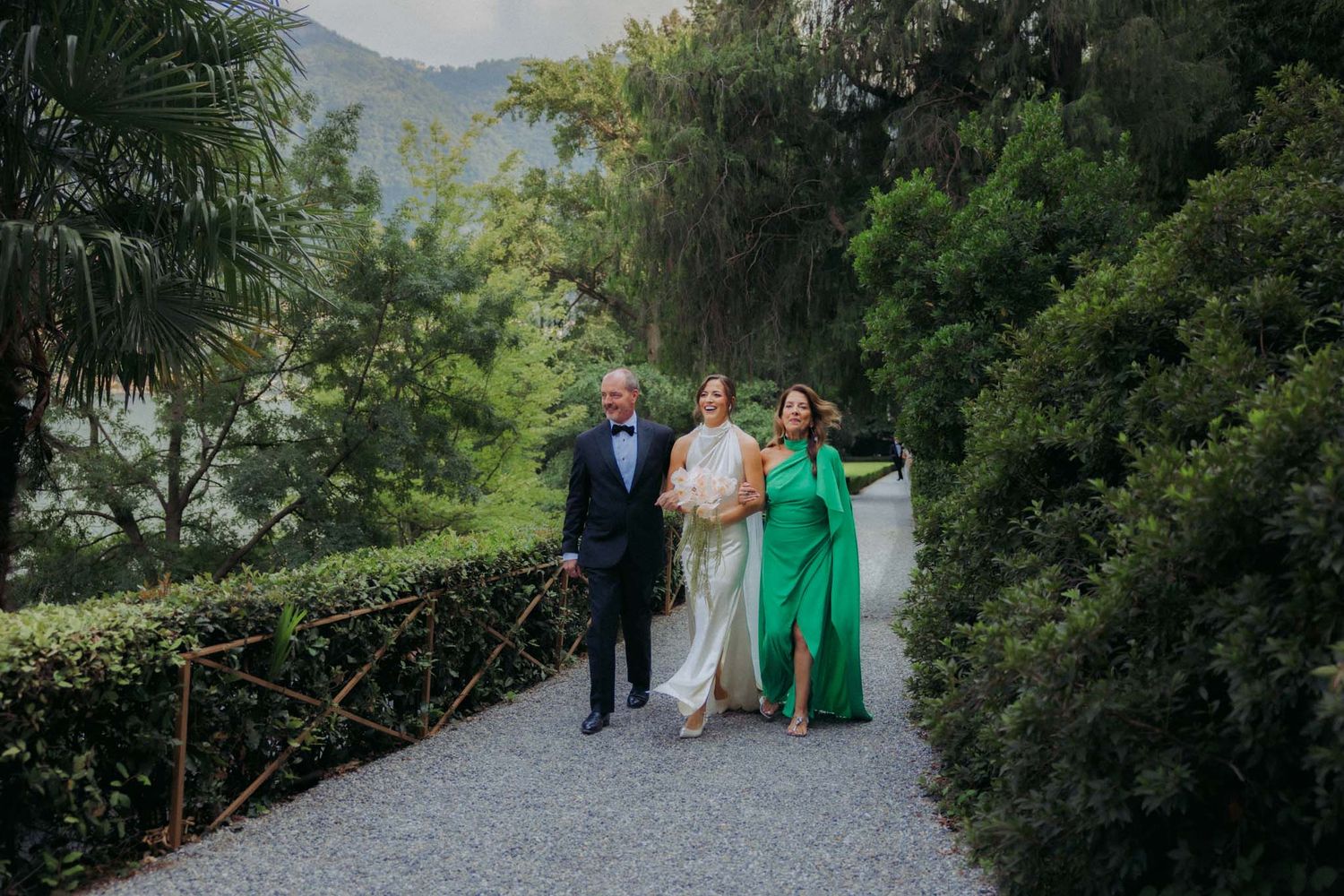 Three people walk arm in arm down a garden path surrounded by lush greenery and mountains in the background.