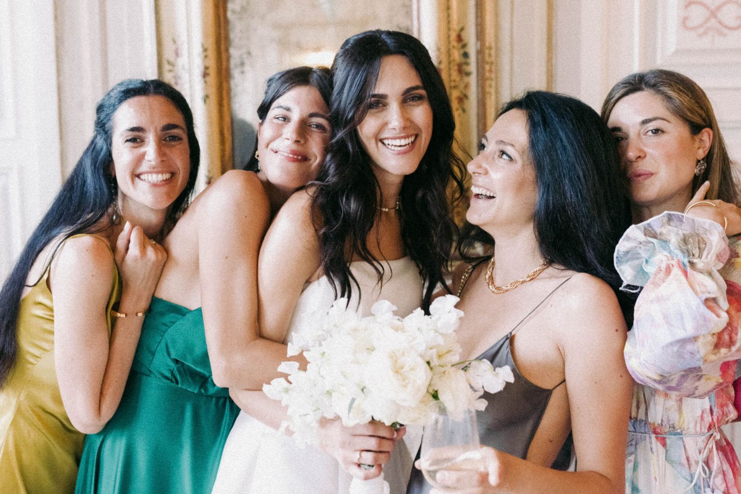 A group of friends in colorful dresses smile and laugh together while holding white flowers at a celebration.