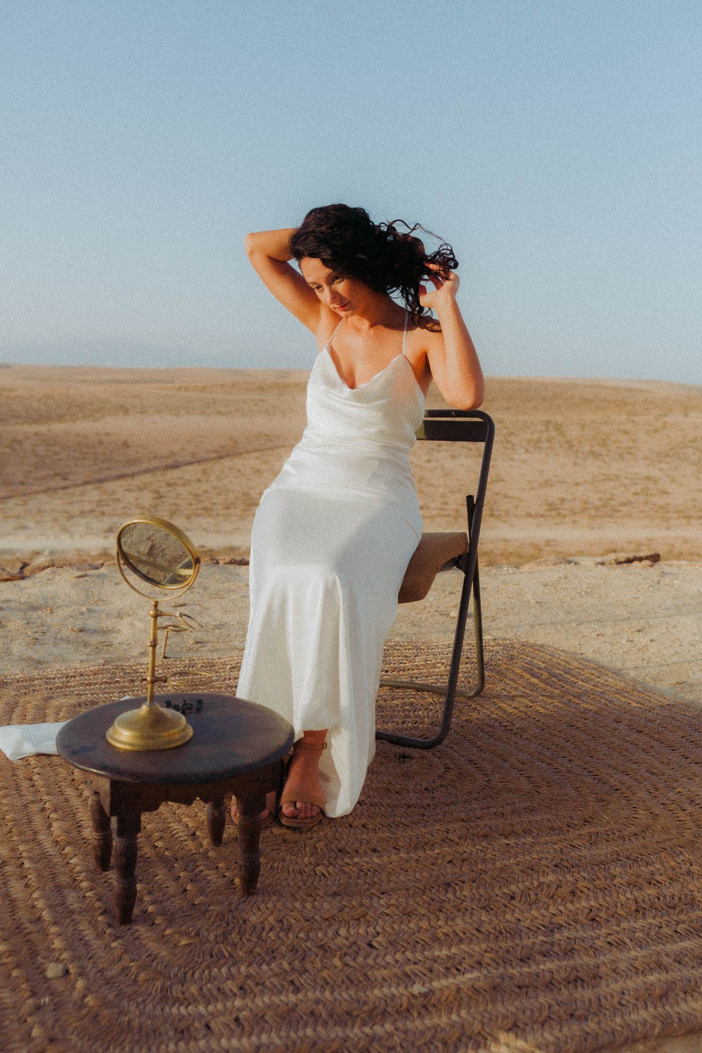 A figure in a flowing white dress poses in a desert landscape with a vintage table and mirror at sunset.