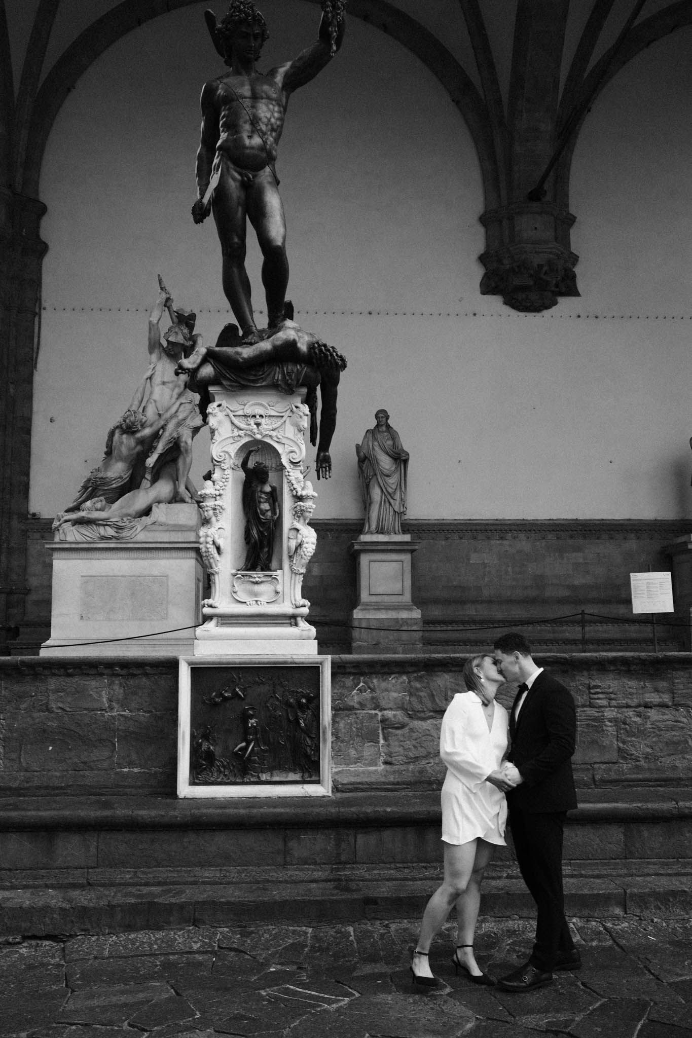 A romantic black and white photo series showing a couple embracing beneath classical statues in an outdoor gallery.
