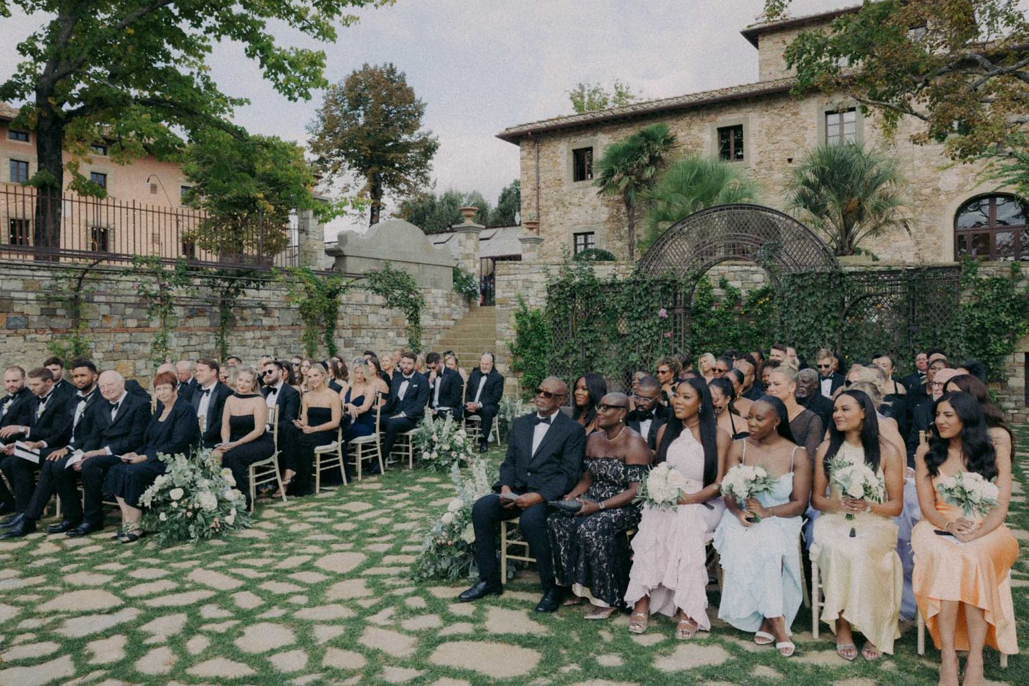Wedding guests gather in an elegant stone courtyard garden for an outdoor ceremony in Italy.