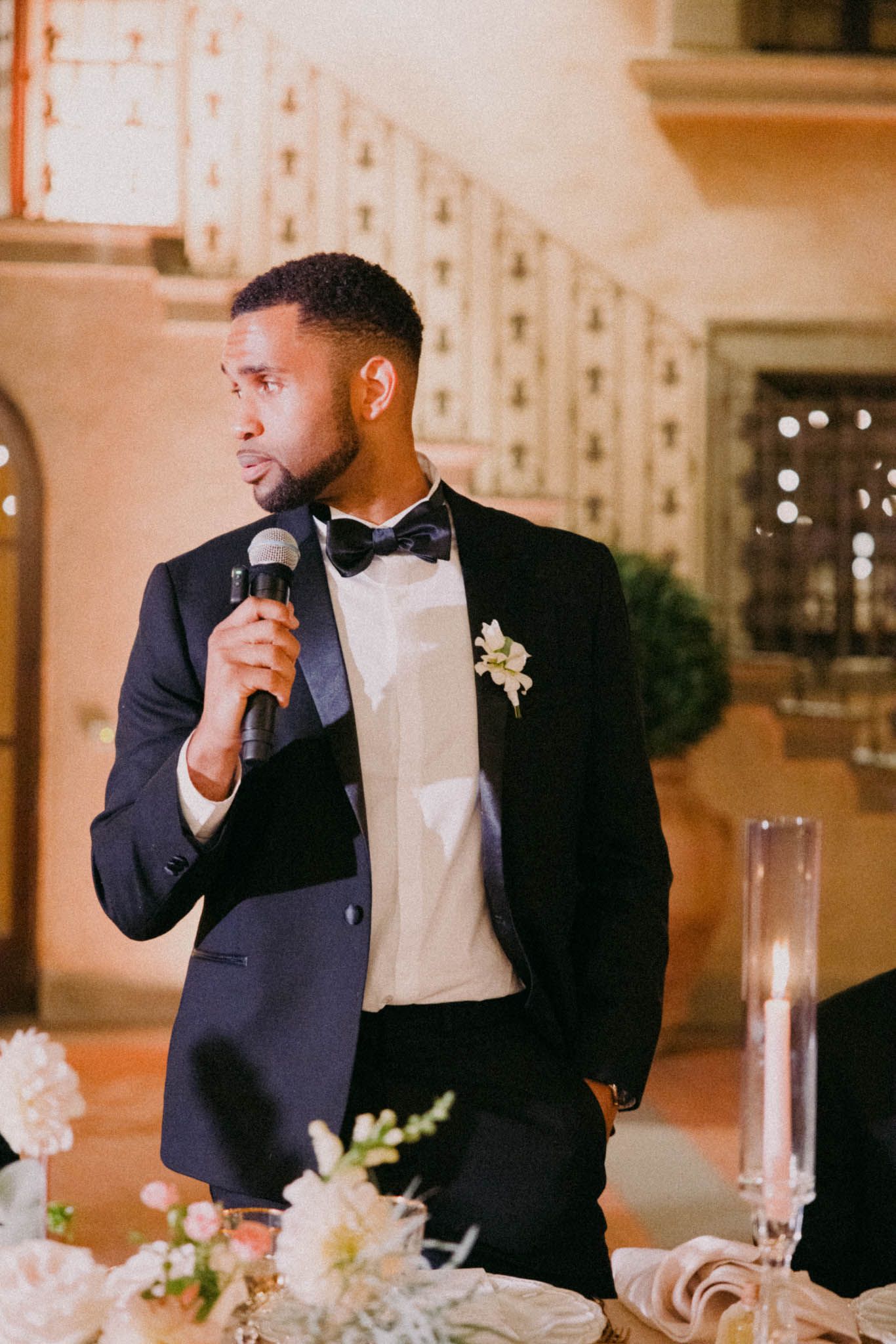 A person in a black tuxedo and bow tie gives a speech at an elegant wedding reception with floral centerpieces.