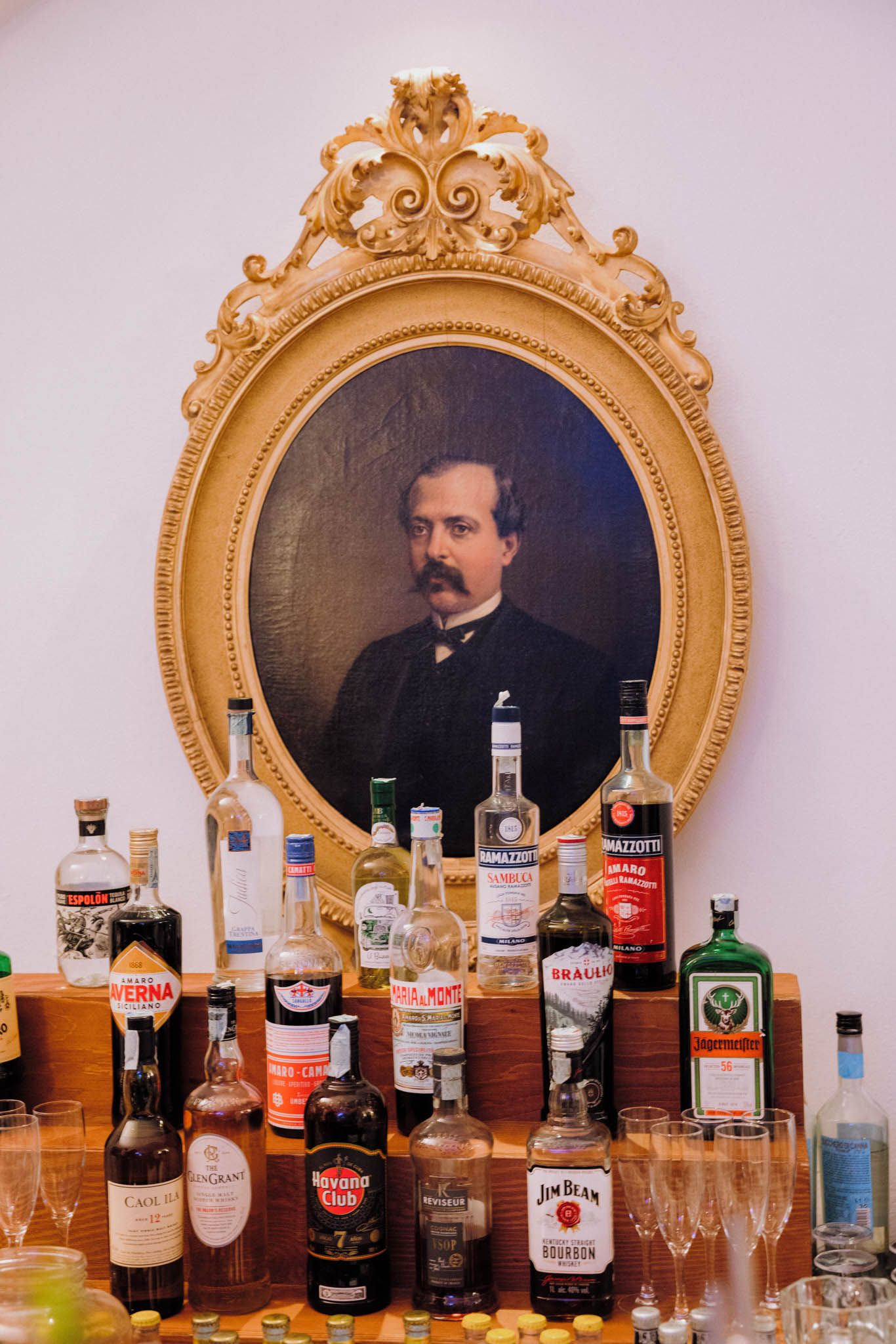 Ornate gold-framed portrait hanging above collection of liquor bottles and glasses arranged on wooden bar display.