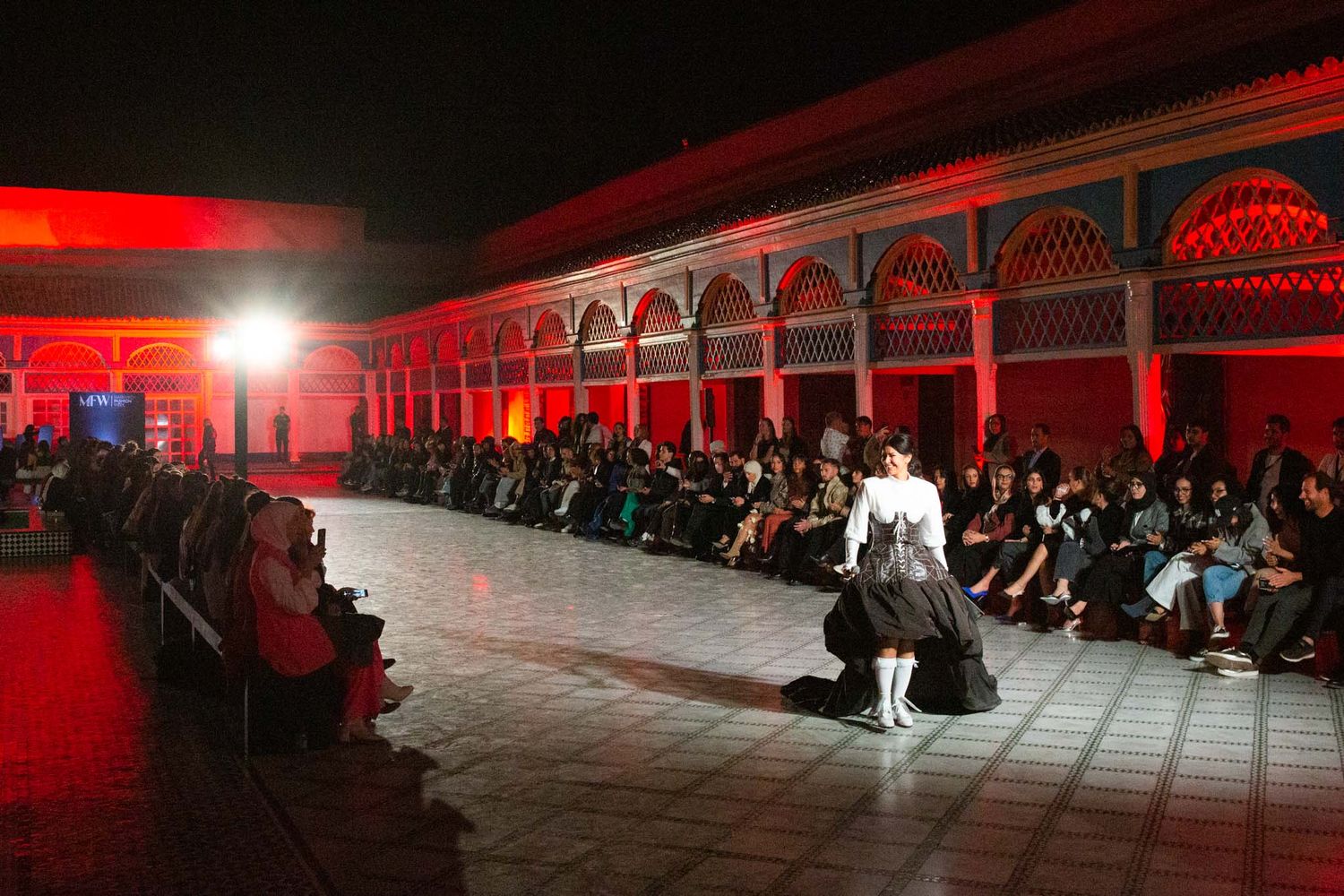 Fashion show runway in historic courtyard illuminated with dramatic red lighting and seated audience.