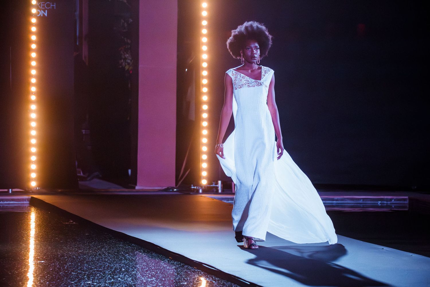 A model walks the runway in a flowing white wedding gown with dramatic lighting on a dark stage.