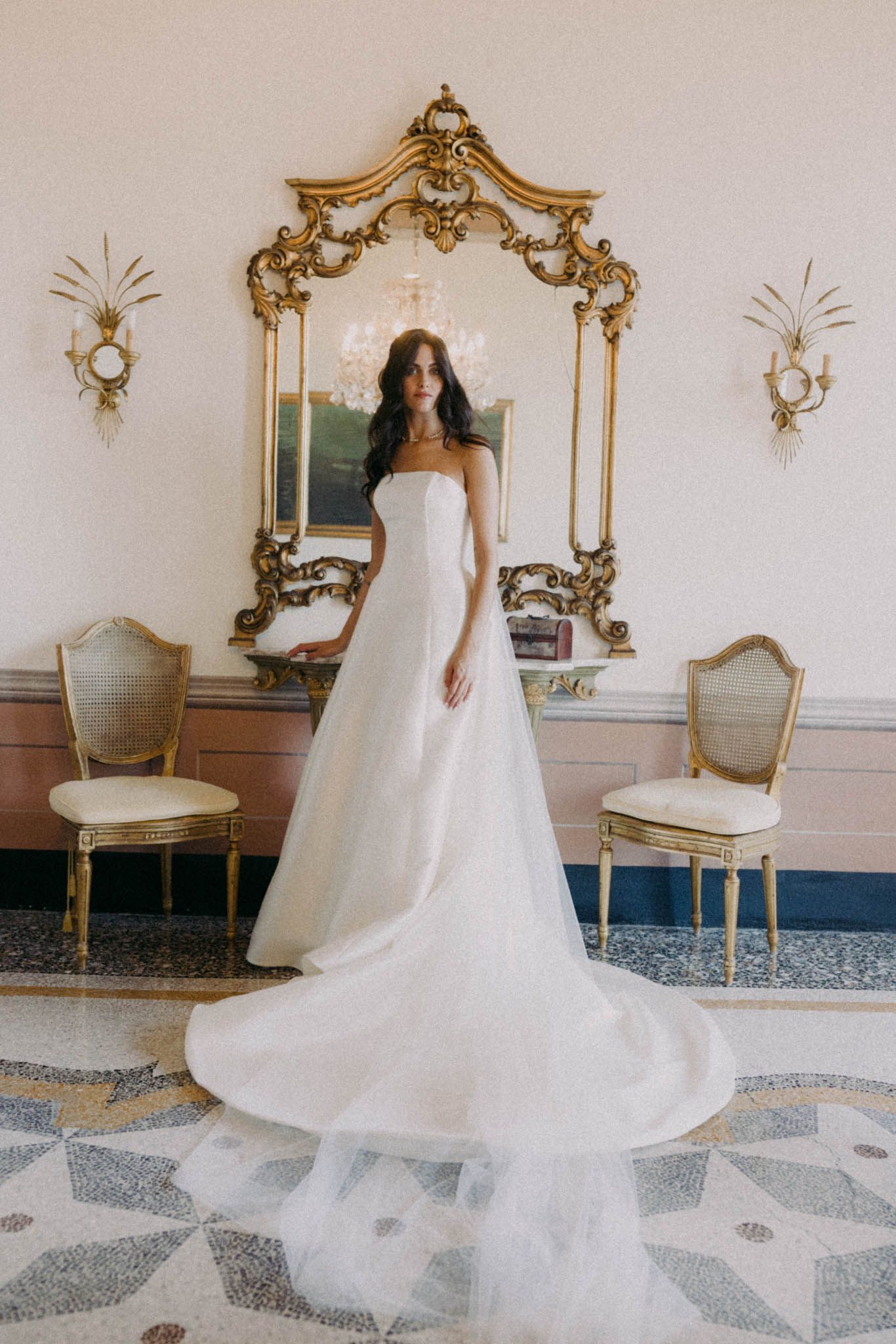Elegant white wedding dress with flowing train photographed in ornate room with gold mirrors and decorative details.