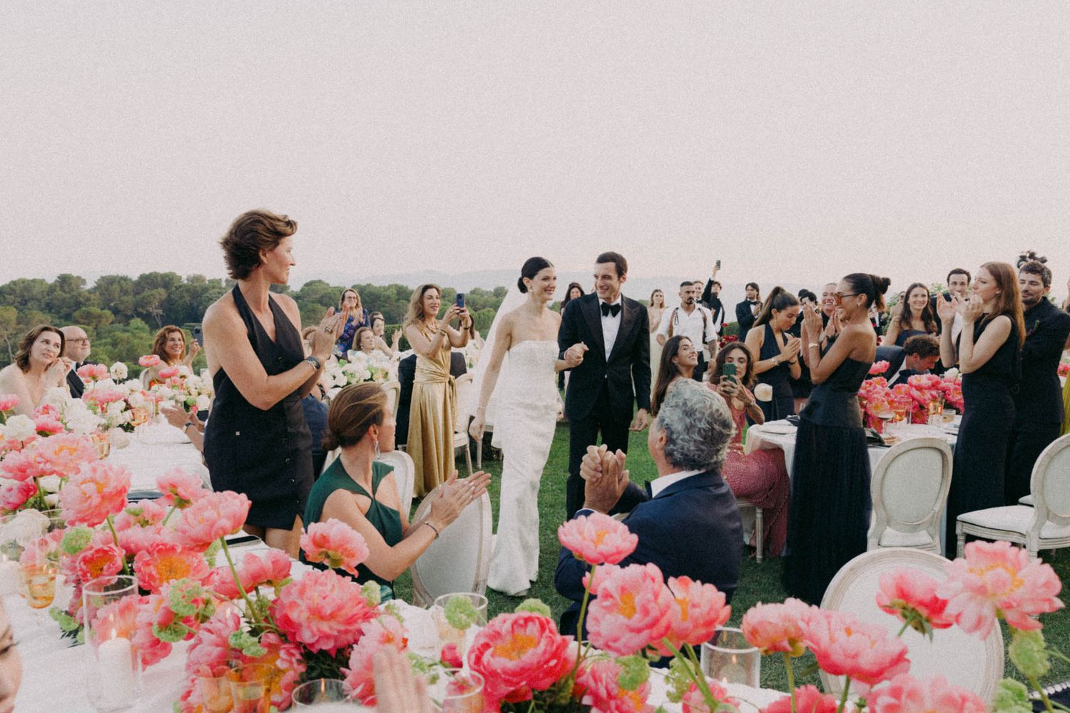 Wedding guests gather around elegant tables decorated with vibrant pink floral centerpieces at an outdoor reception.