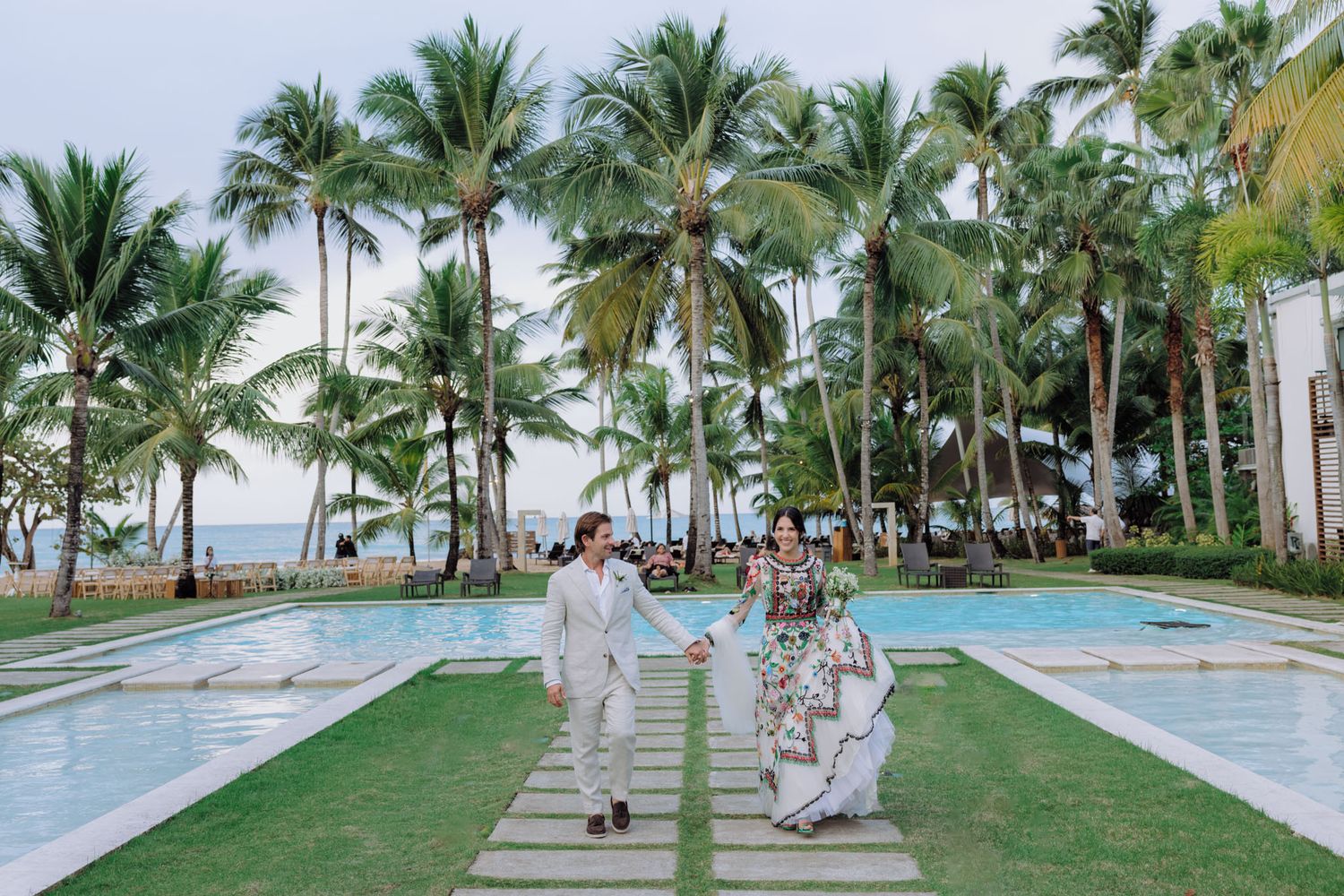 Palm trees line a beachfront walkway with people strolling along the tropical oceanside path.