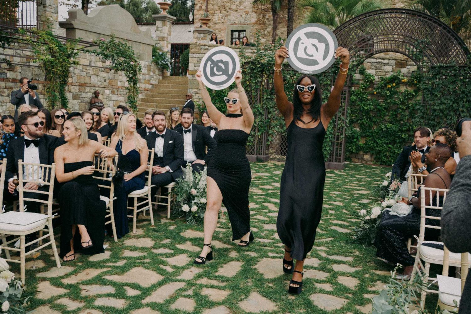 A group of people celebrate at an outdoor wedding reception in a garden courtyard setting with stone walls and greenery.