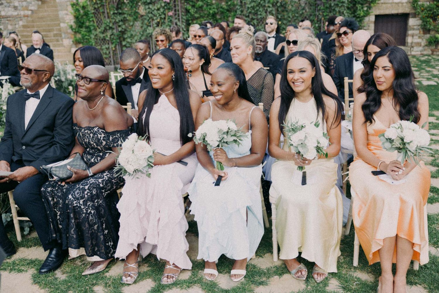Wedding guests seated in formal attire at an outdoor garden ceremony holding white bouquets.