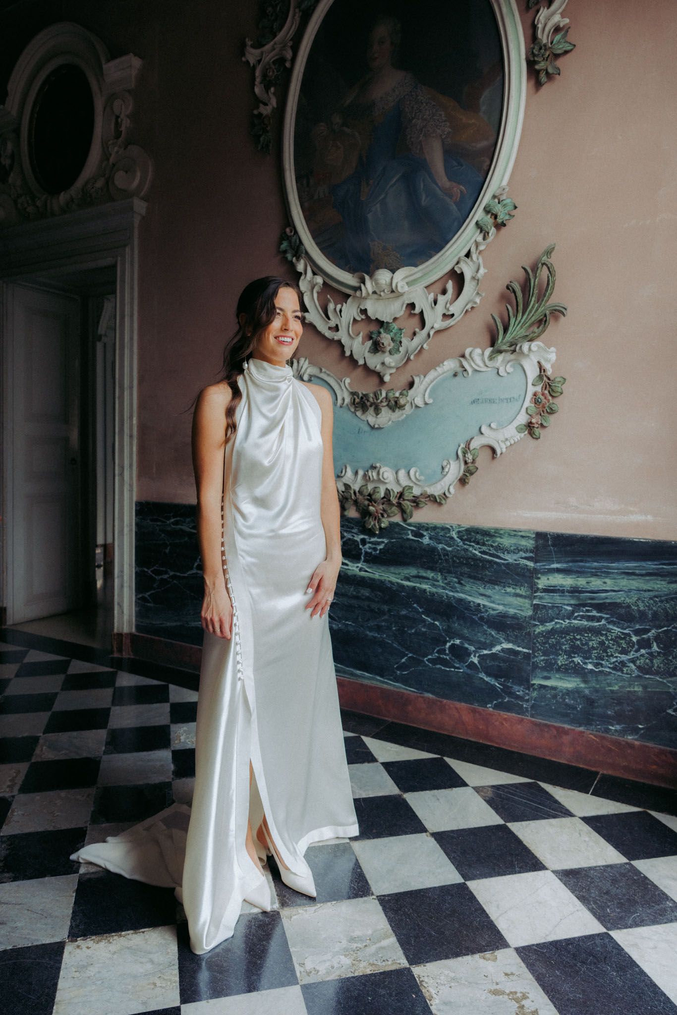 A person in a flowing white dress poses on a black and white checkered floor against an ornate wall with vintage decor.