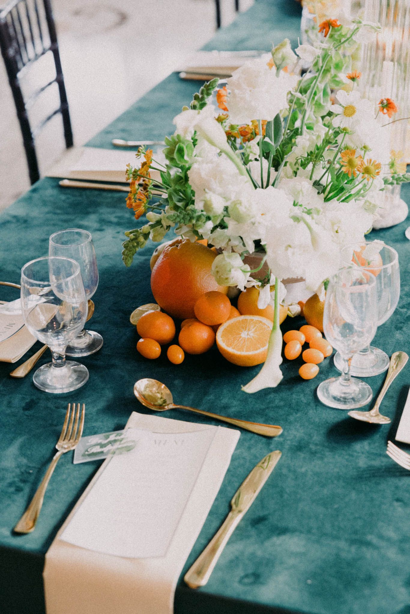 Elegant table setting with white flowers, oranges, and glassware on dark teal velvet tablecloth.