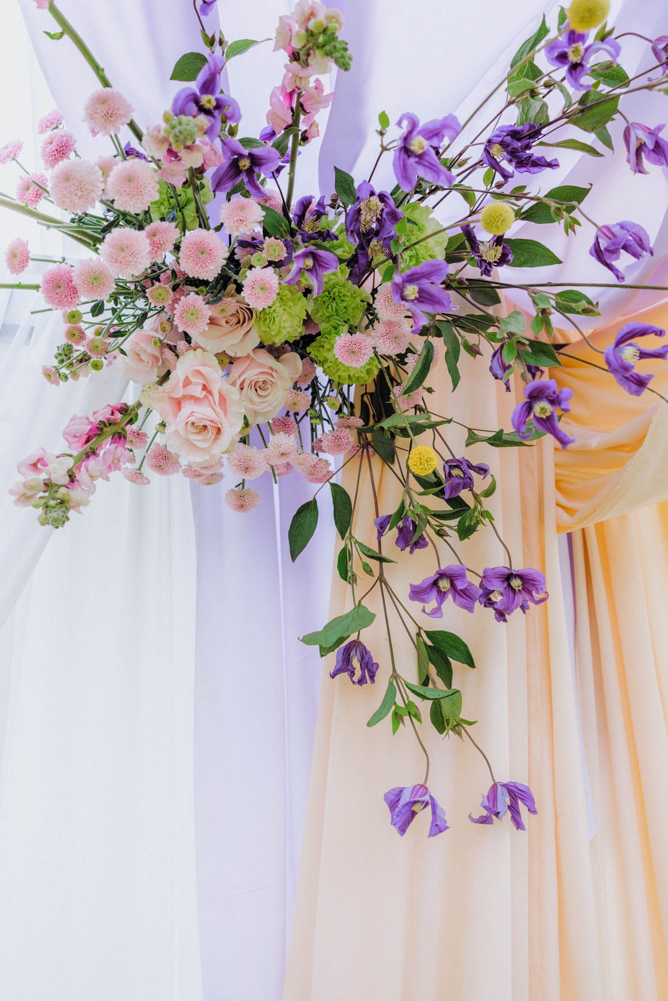 Elegant floral arch decoration featuring pink roses, purple iris flowers and greenery against white and peach drapery.