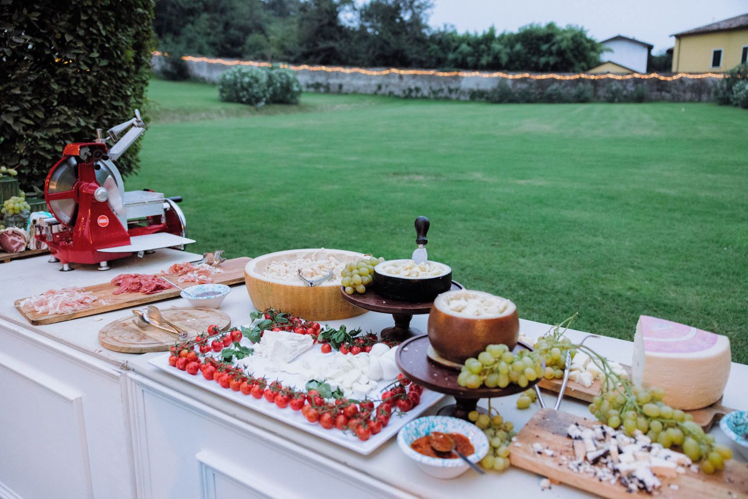 An elegant outdoor catering spread features assorted cheeses, fresh fruits, and appetizers on white serving platters.