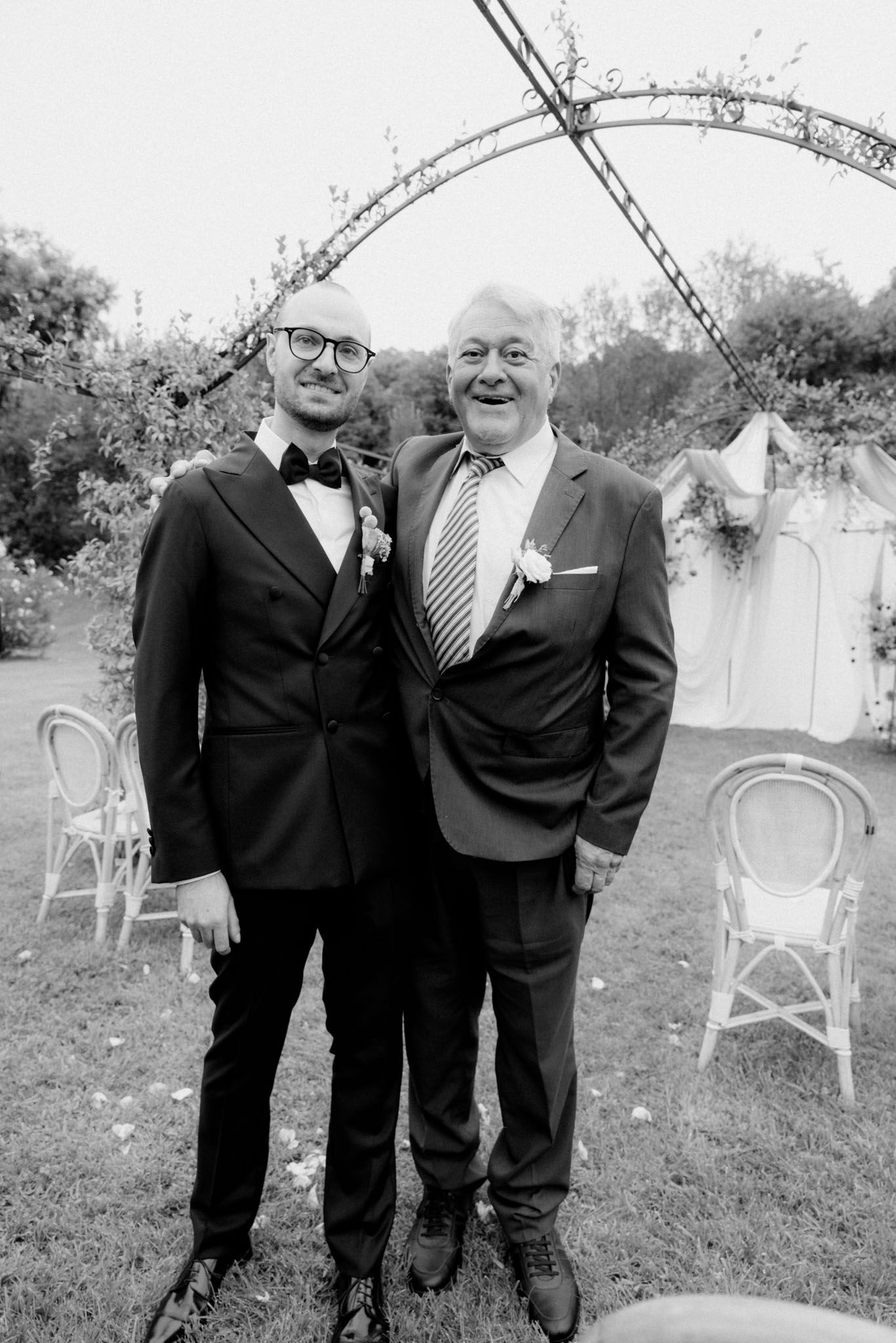 Two men in formal black suits stand together on grass with white garden chairs visible in the background.