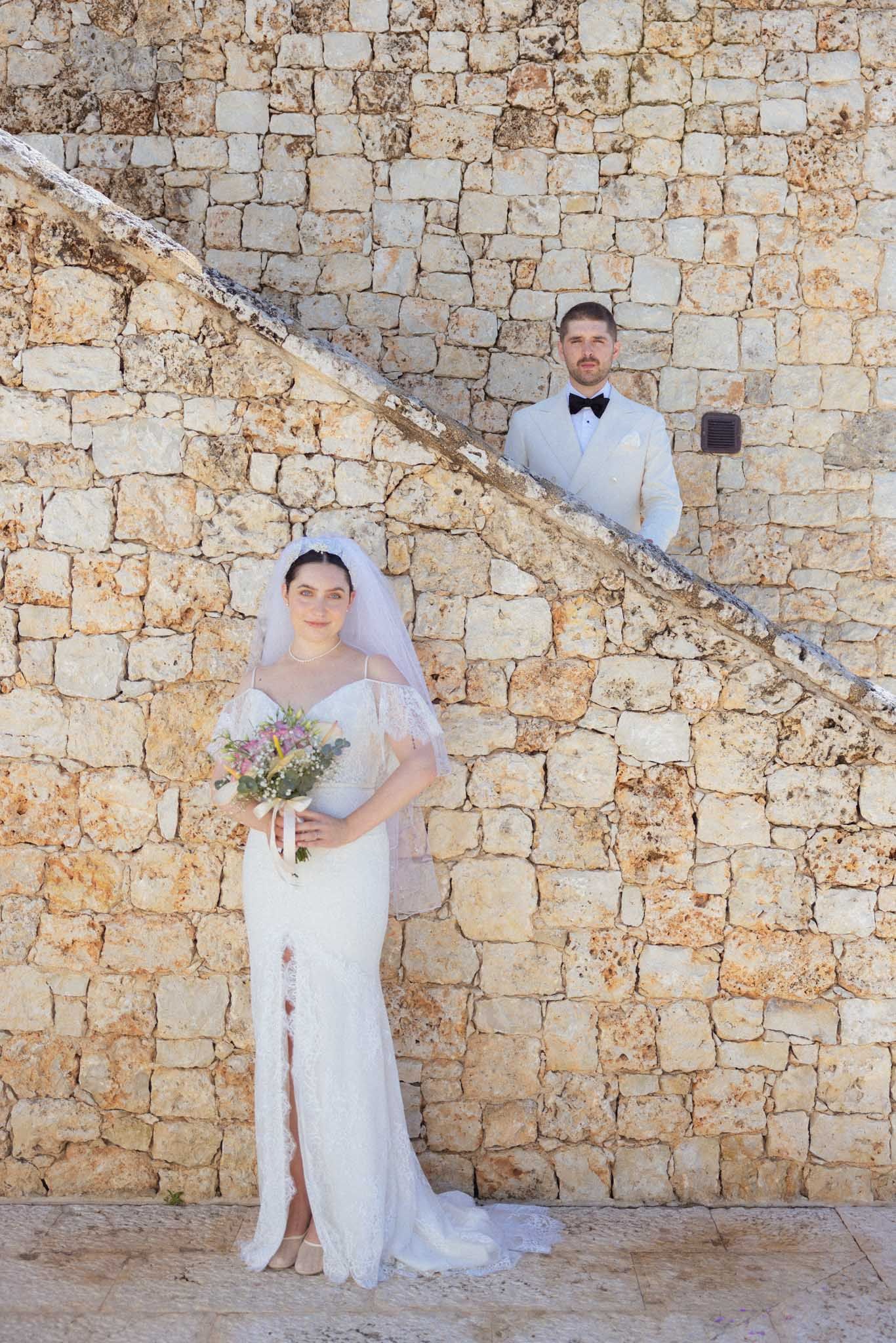Wedding couple poses against rustic stone wall in series of romantic portraits wearing elegant white attire.