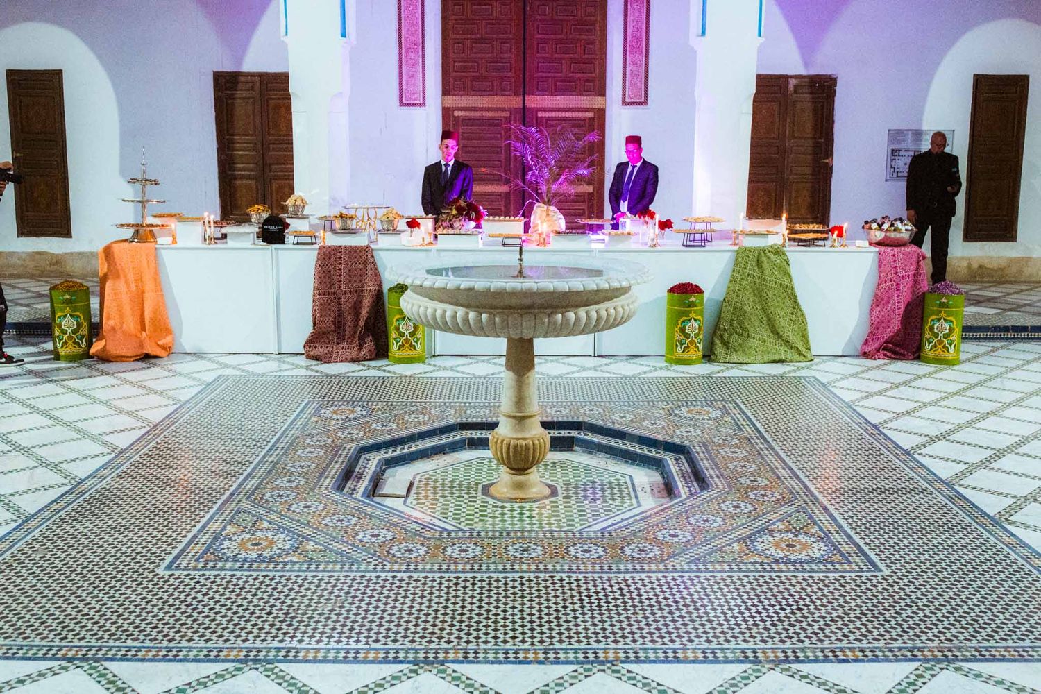 Interior courtyard of a Moroccan palace featuring ornate tile patterns and fountains illuminated in purple and pink light.