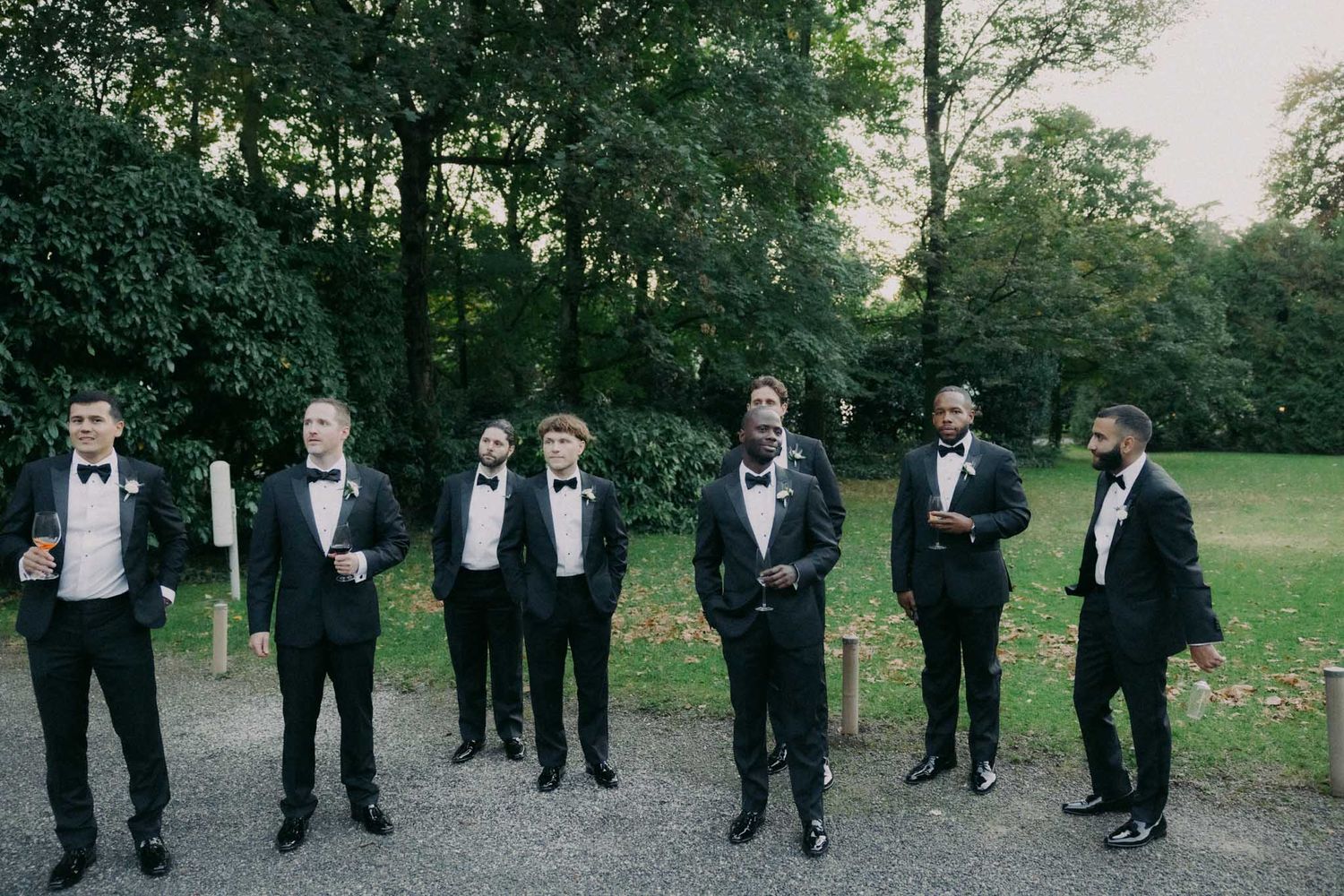 A formal wedding party wearing black tuxedos poses outdoors in a garden setting.