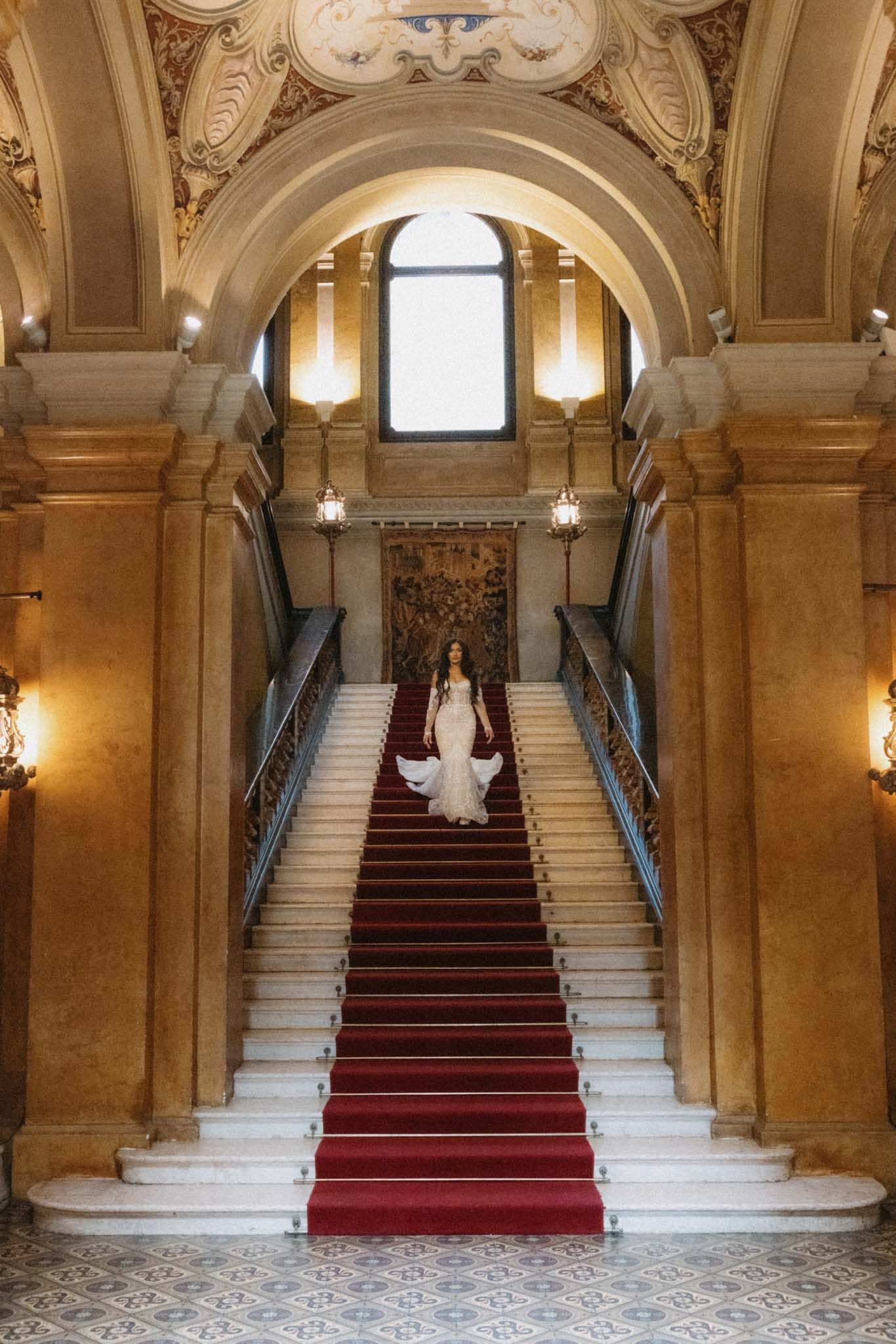 Elegant grand staircase with red carpet runner and ornate stone architecture in historic palace interior.