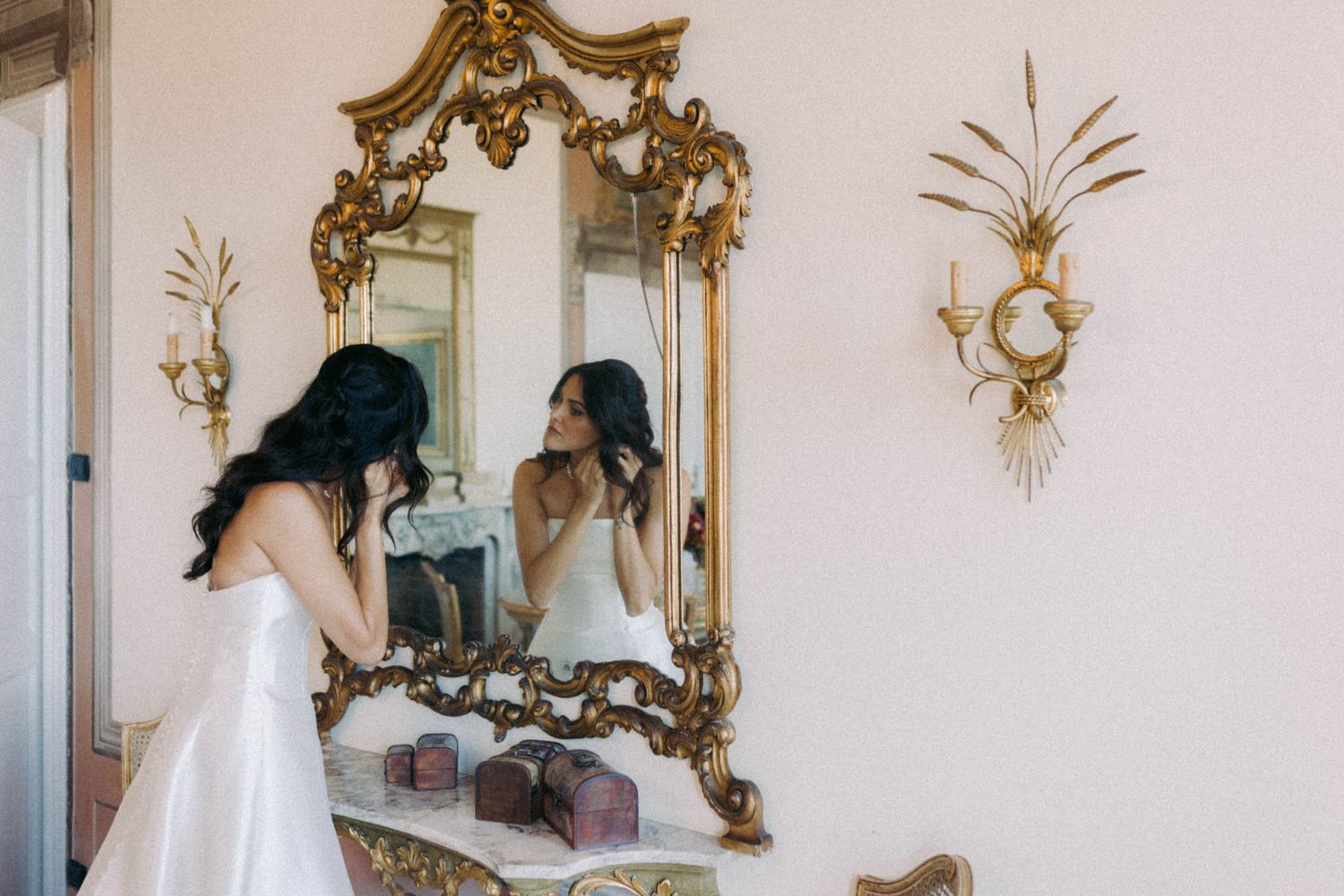 A woman in a white dress looks at her reflection in an ornate gold mirror with decorative wall sconces.