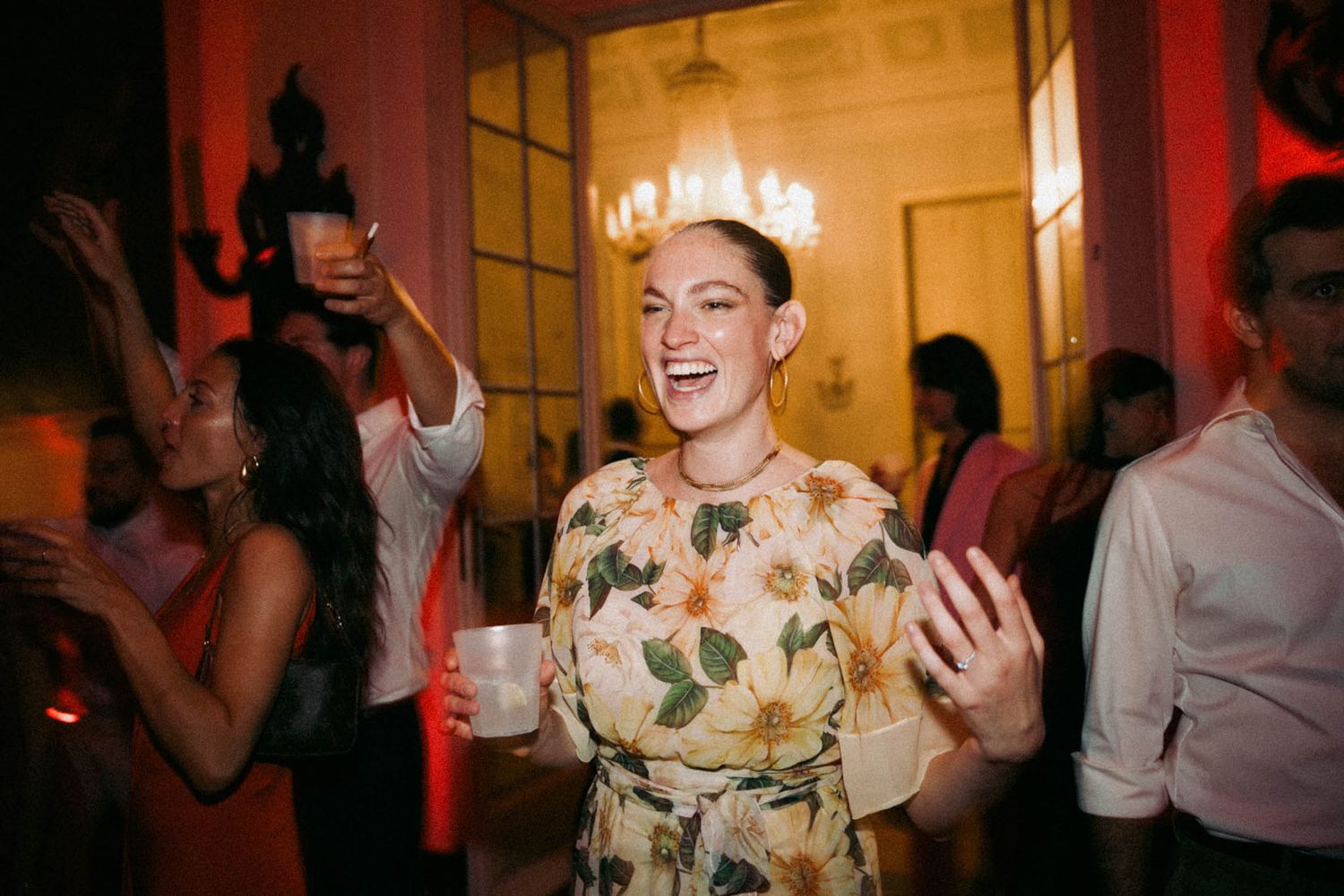 Person in floral dress dances joyfully at a party with glowing lights and other guests in the background.