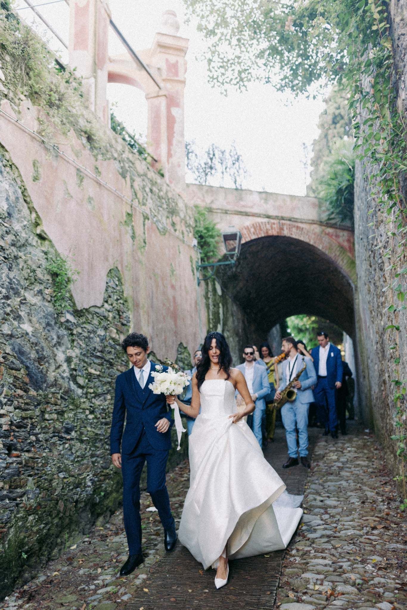 Wedding couple walks through a romantic stone archway tunnel with their wedding party following behind them.