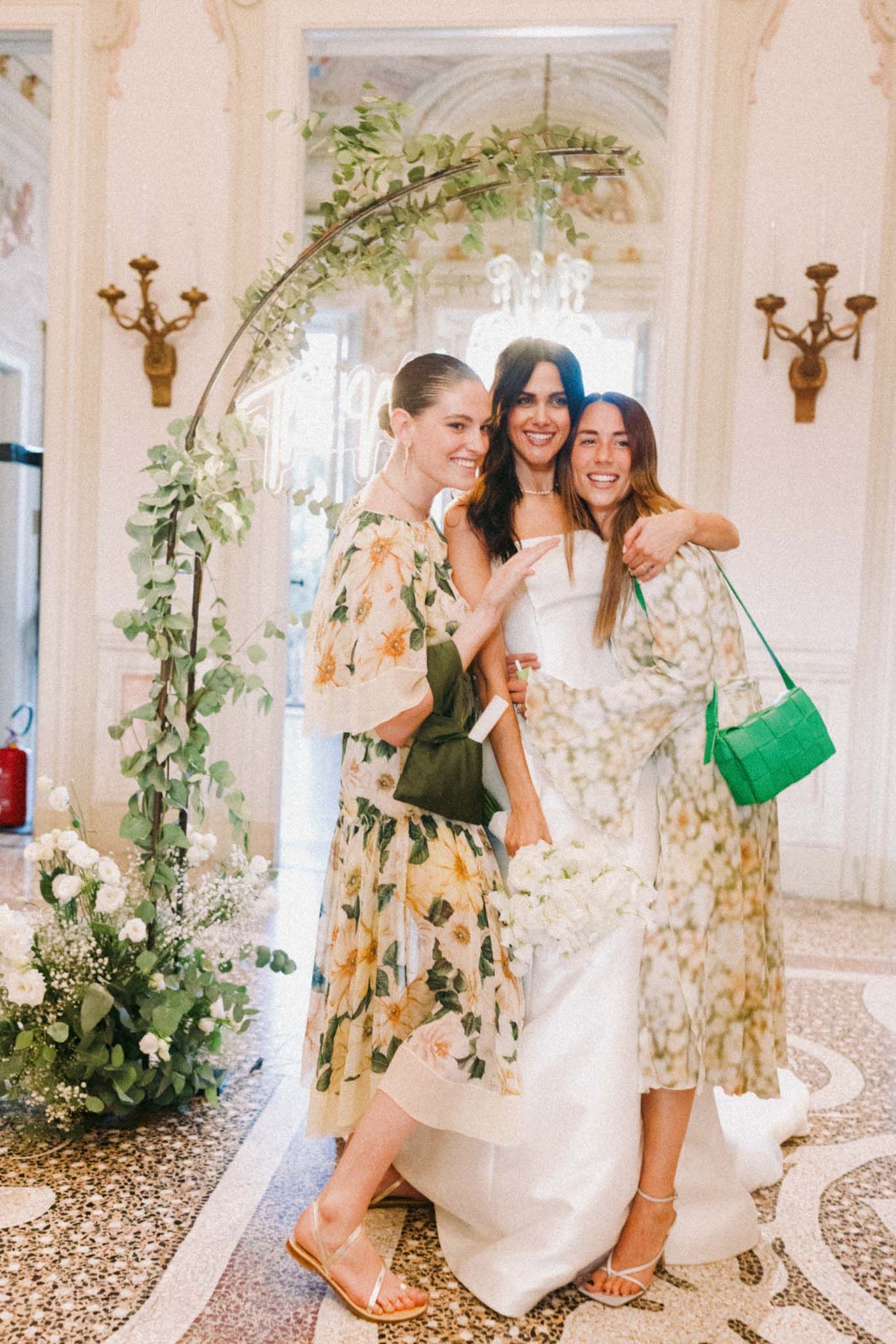 Three women in floral dresses laugh and dance together in an elegant white room decorated with greenery and candelabras.