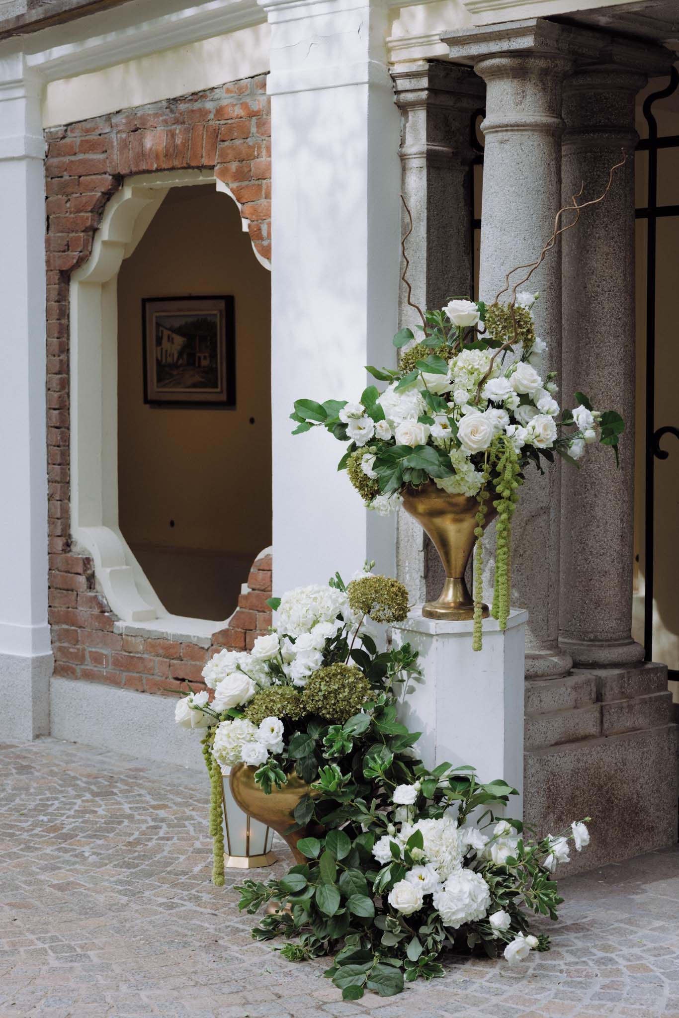 Elegant white floral arrangements in gold vases adorn a brick and stone archway entrance with classic architectural details.