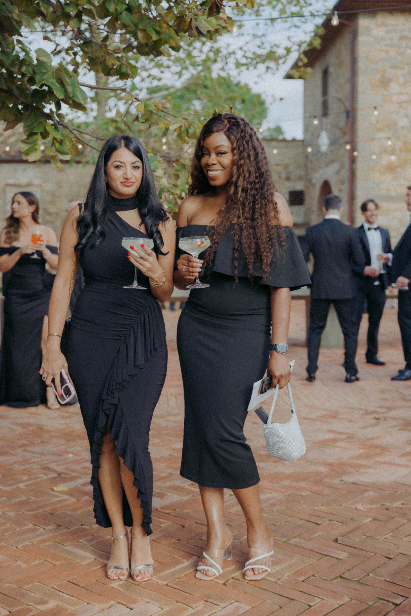 Wedding guests in black dresses socialize and mingle on a rustic stone patio during an outdoor celebration.