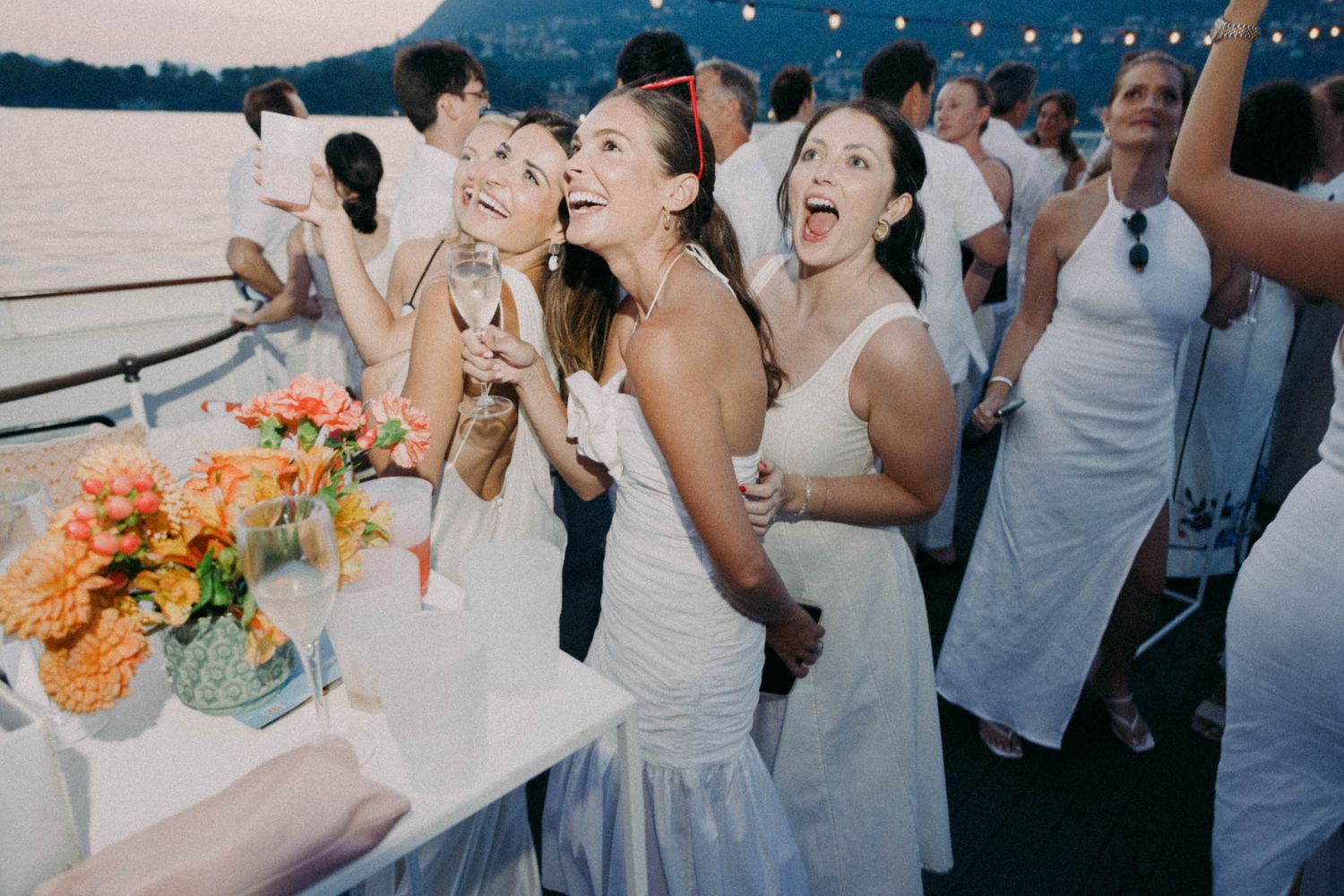 Wedding guests celebrate joyfully on a boat deck at sunset with floral centerpieces and string lights overhead.