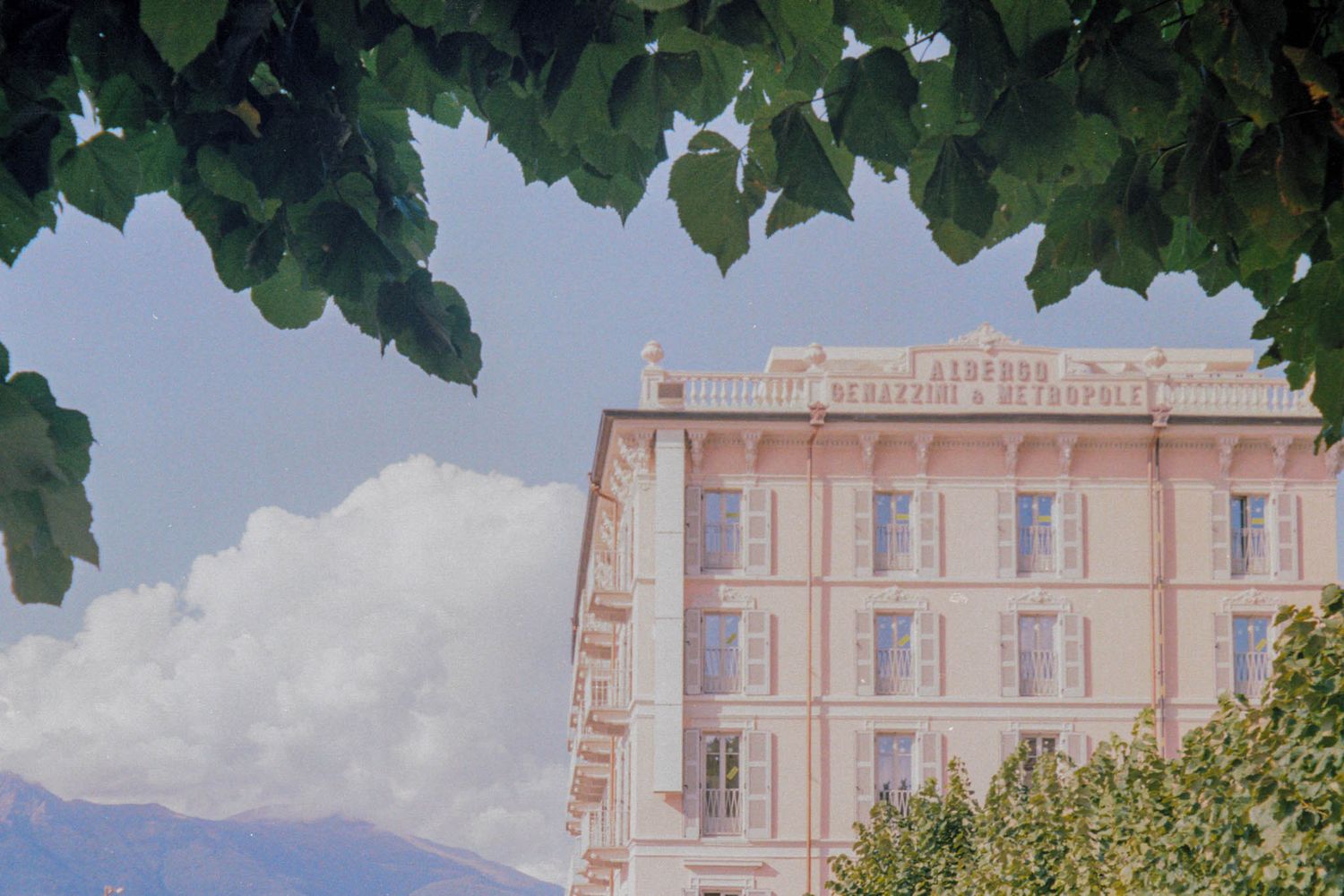 Elegant pink-hued hotel building with traditional French architecture and leafy green trees framing the view.