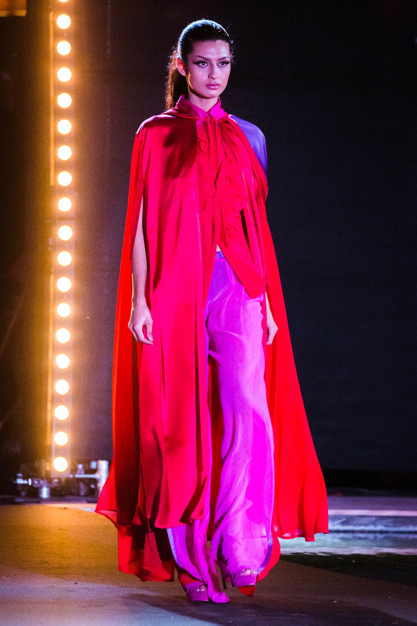 Model walks the runway in a flowing bright red gown with dramatic cape sleeves under stage lighting.
