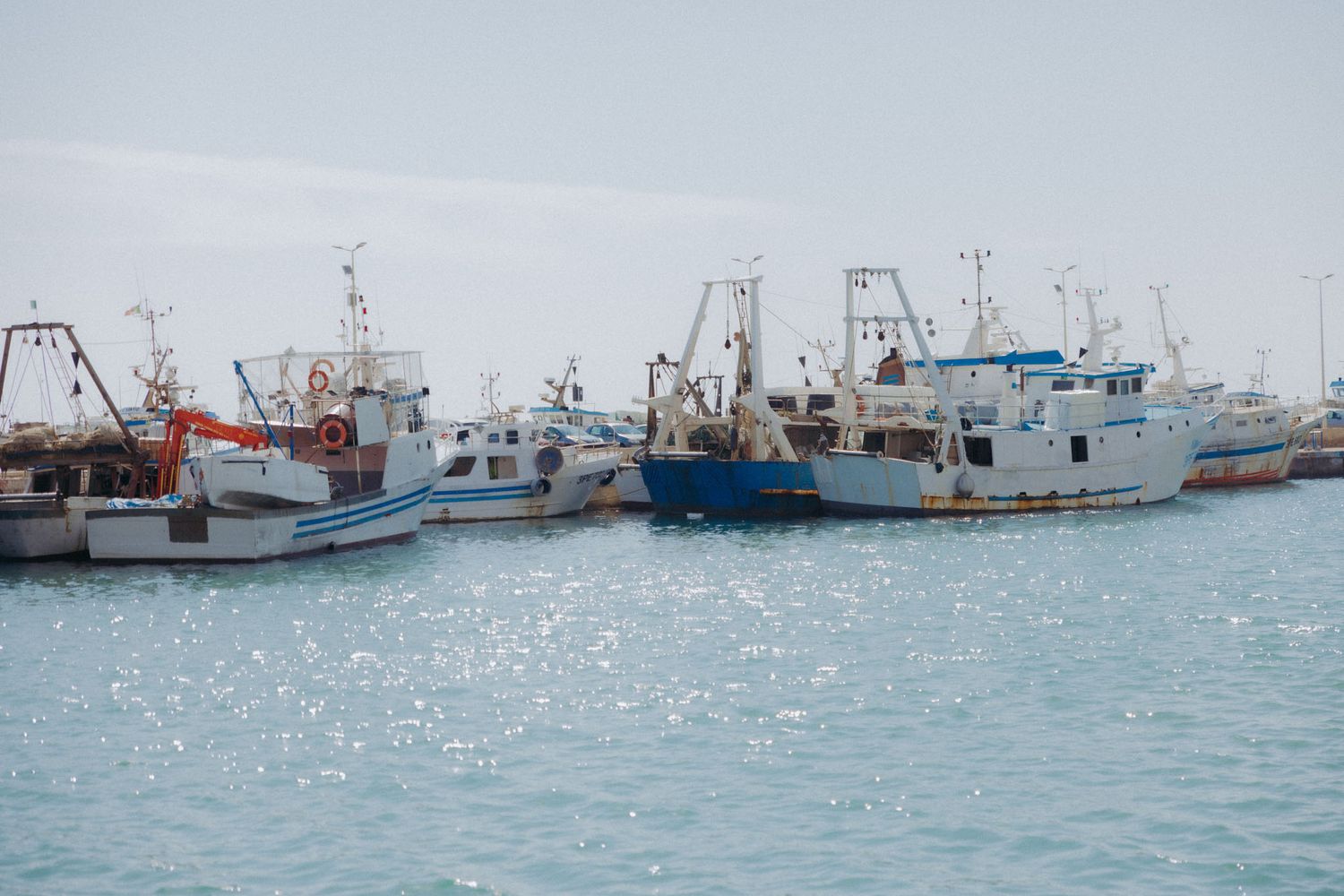 Multiple fishing boats and vessels docked at a marina with calm blue waters and cloudy skies in the background.