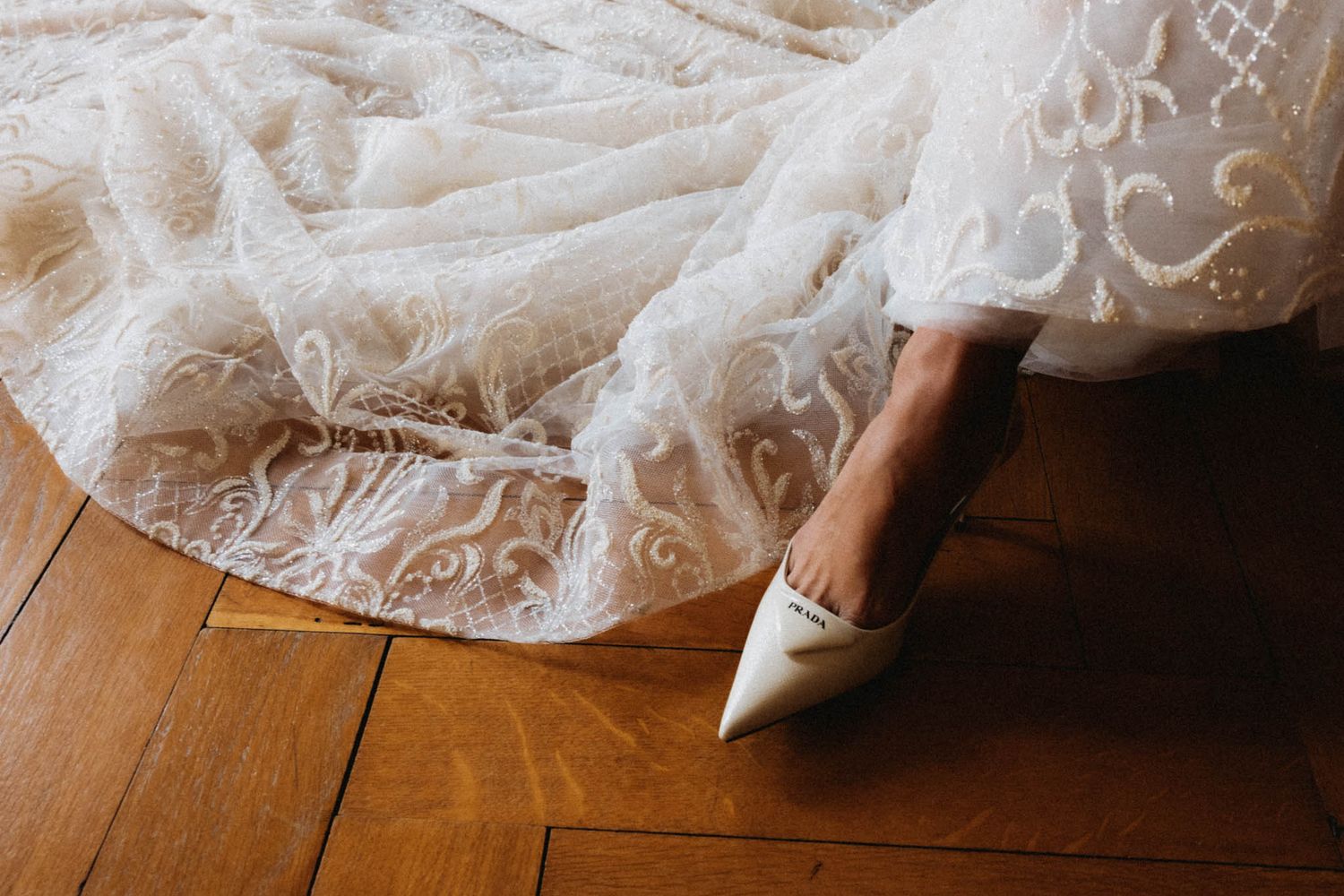 Delicate lace wedding dress details cascade across a rustic wooden floor in soft natural lighting.