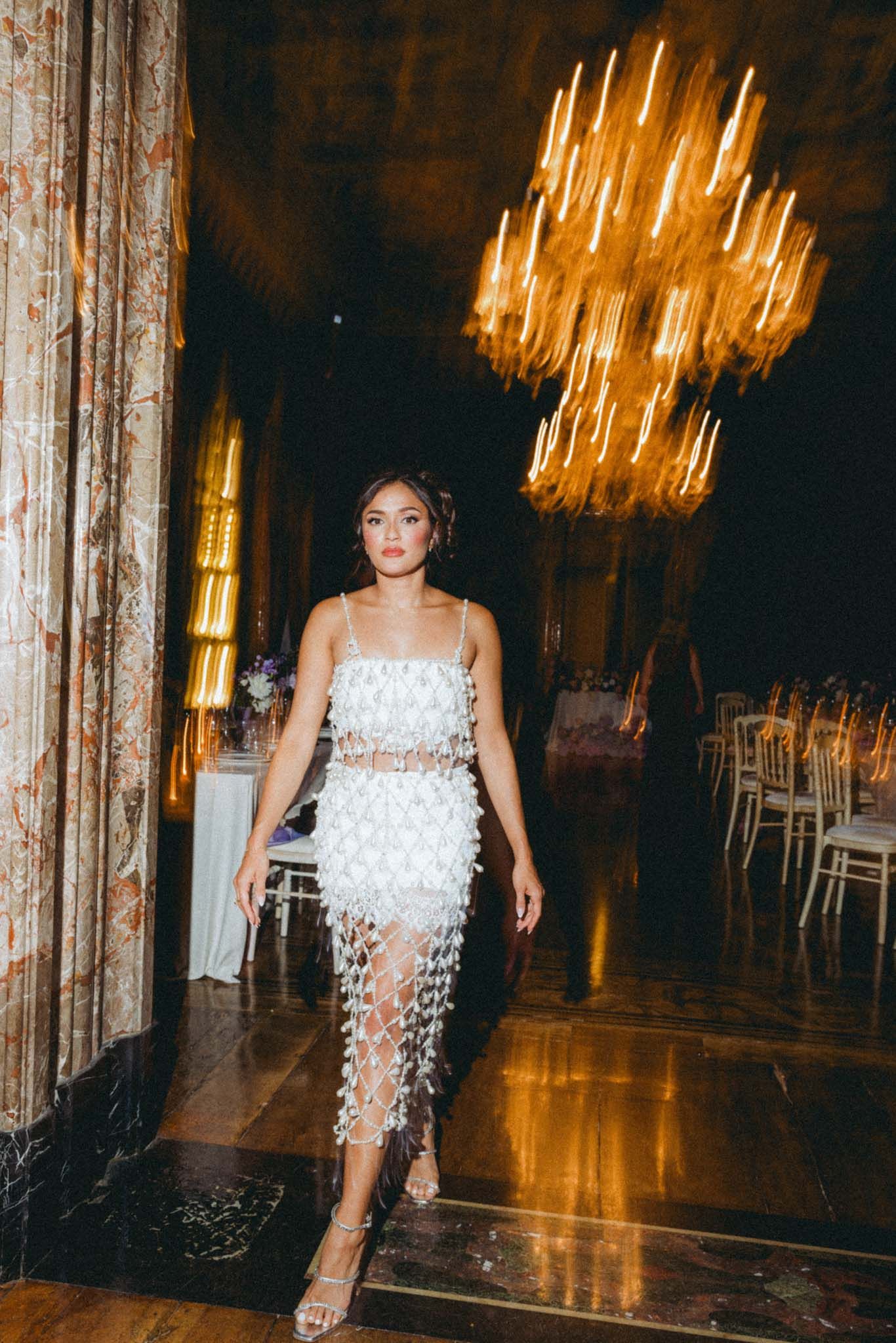 A figure in an elegant white dress adorned with delicate feathers stands in an opulent ballroom with crystal chandeliers.