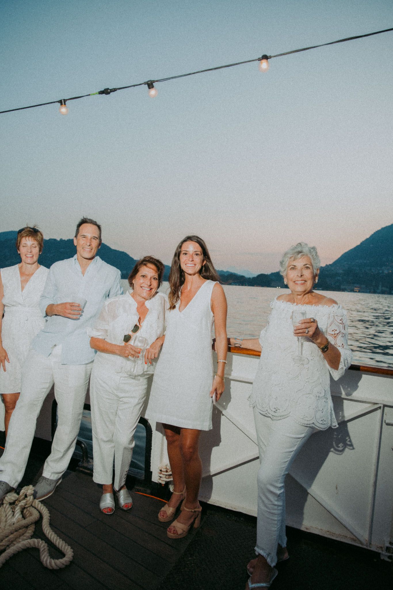 A group of people in white outfits pose together on a boat deck at dusk with string lights and mountains in the background.