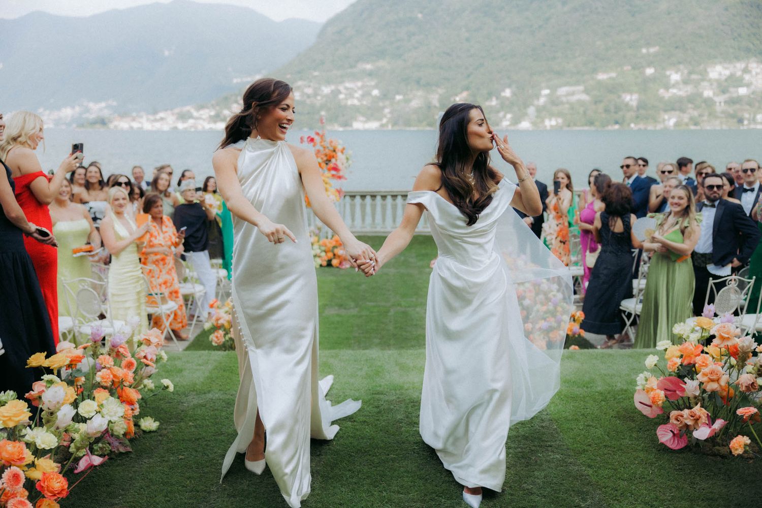 Wedding party members in white dresses dance joyfully on a lawn overlooking a scenic mountain lake landscape.