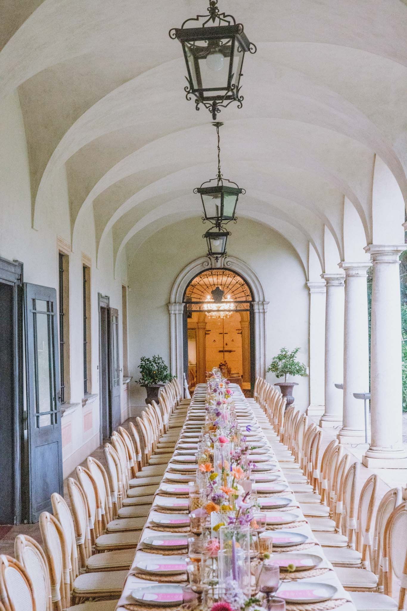 Elegant dining setup in a historic colonnade with white arches, featuring long tables set with pink and white decor.