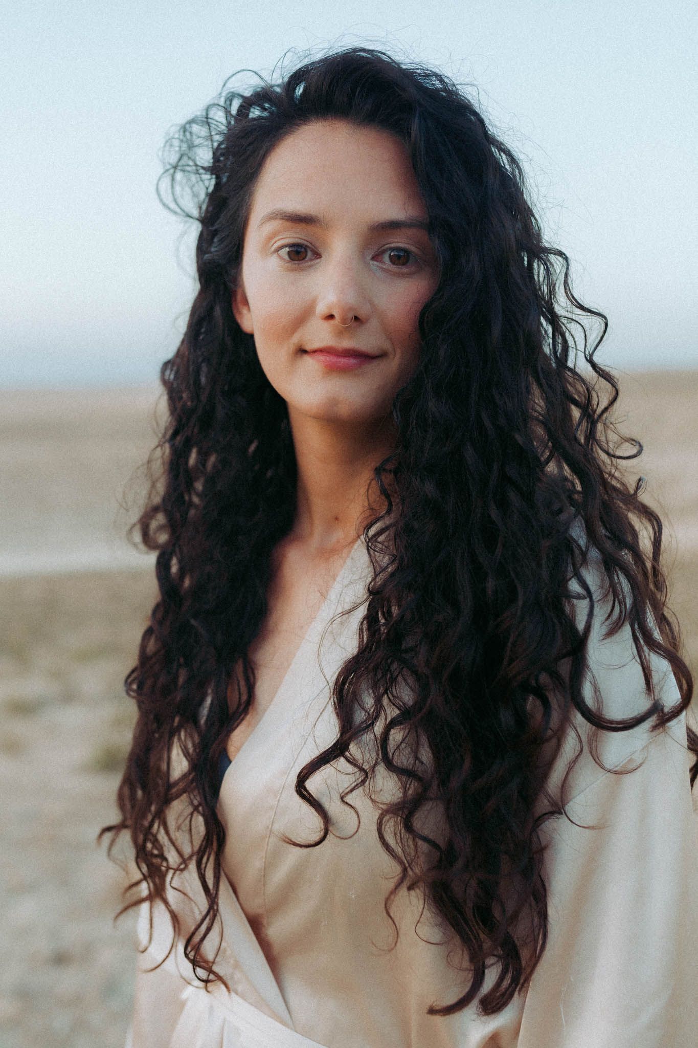 Portrait of someone with long dark curly hair wearing a cream colored top against a light background outdoors.