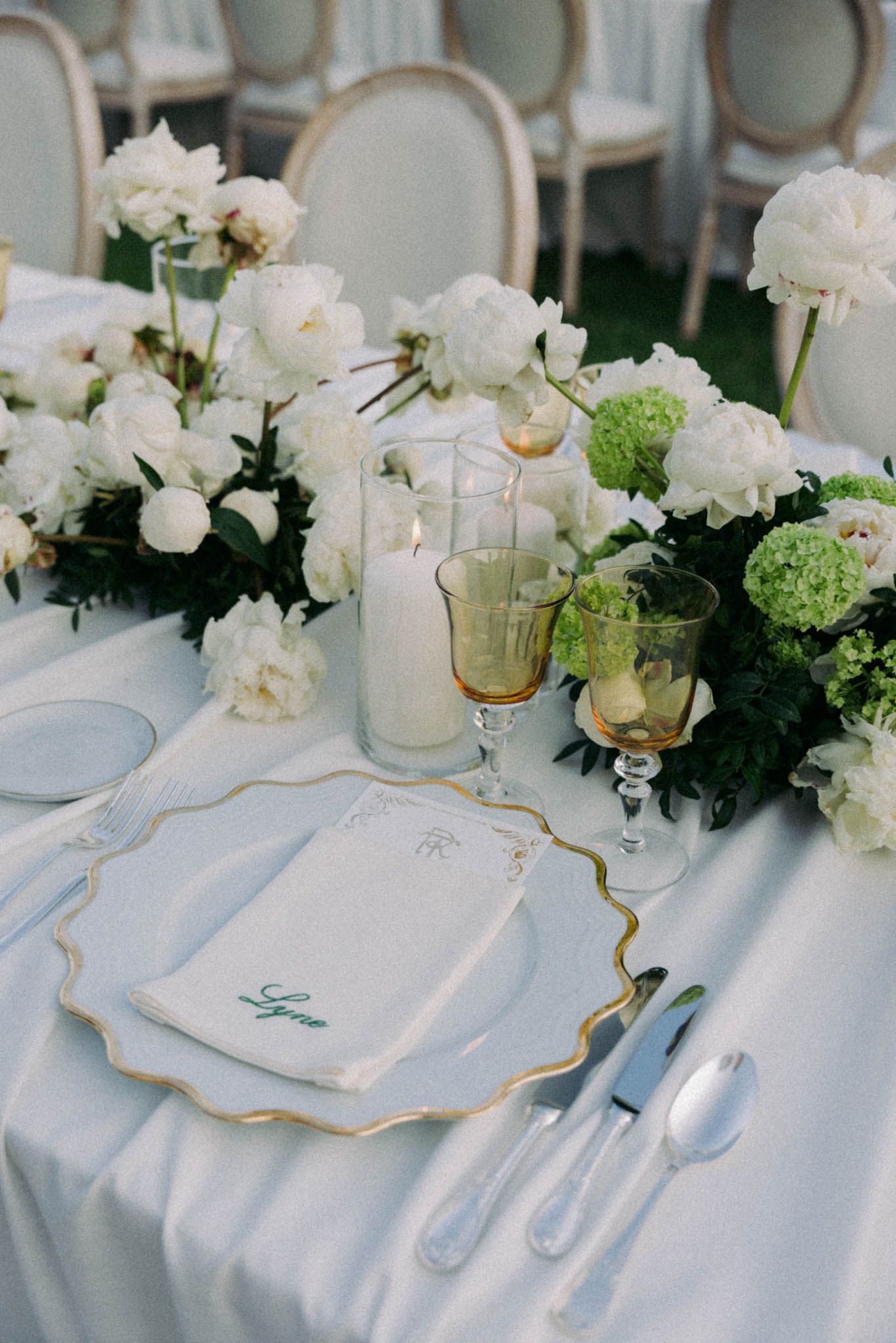 Elegant wedding table setting with white floral centerpieces, gold-rimmed plates and delicate place cards on white linens.