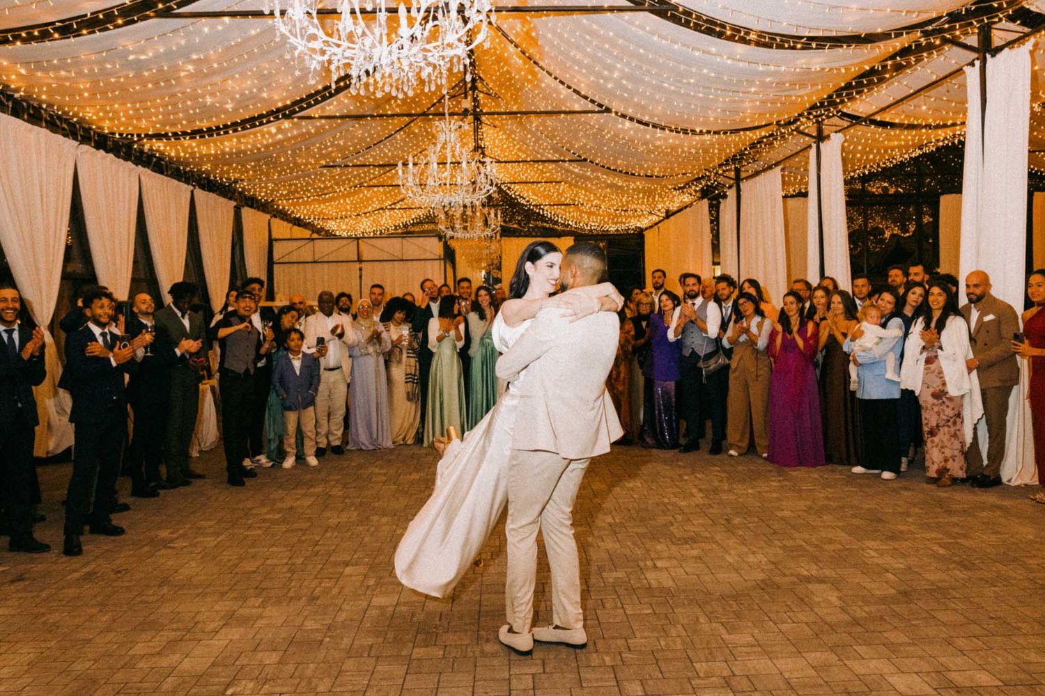 Wedding reception dance floor under twinkle lights and fabric draping with guests gathered around the perimeter.