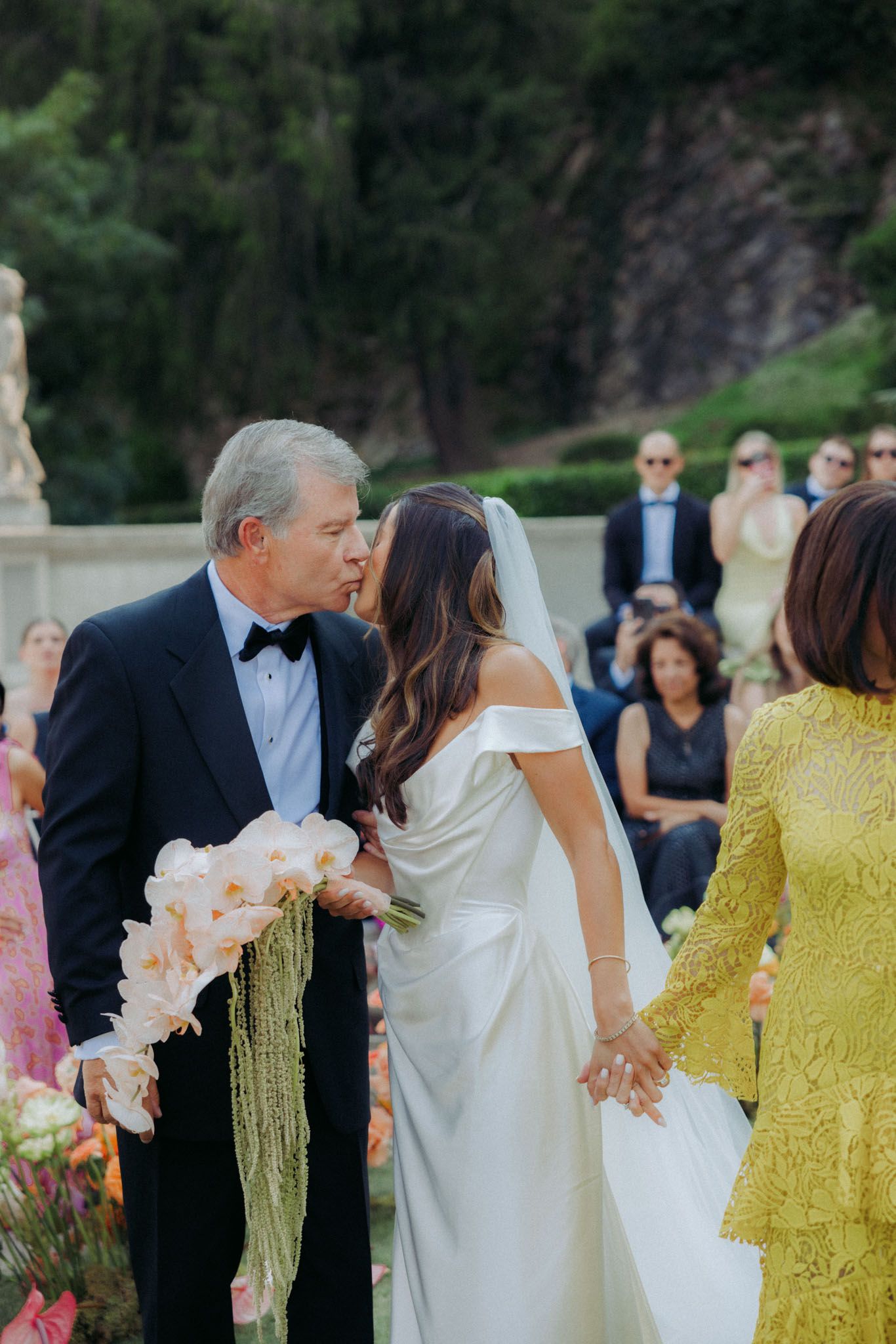 A wedding ceremony processional with attendants in yellow and white dresses walking down an outdoor path.
