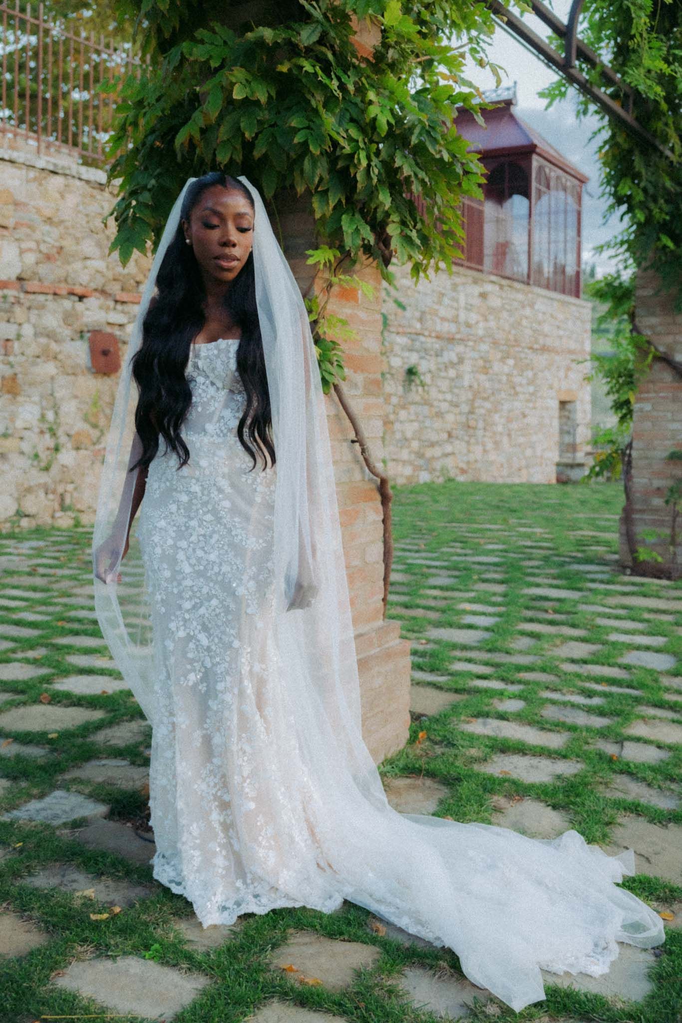 A bride in a stunning white lace wedding gown with a flowing veil poses against a rustic stone wall and greenery backdrop.