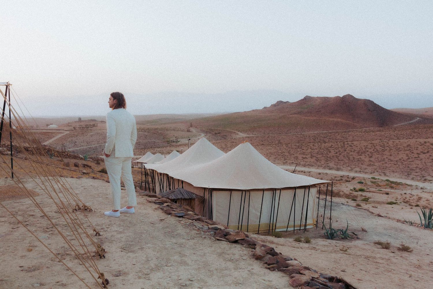 A figure in white stands on a desert cliff overlooking a vast mountainous landscape at sunset.