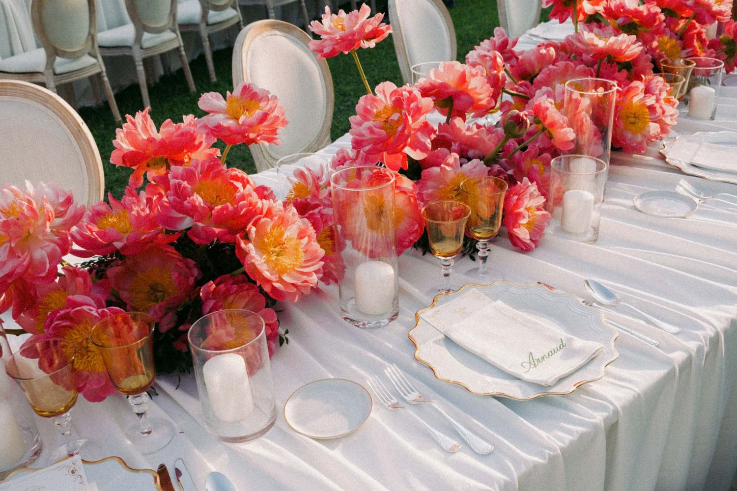 Elegant table setting with vibrant coral and pink floral centerpieces running along a white tablecloth at an event.