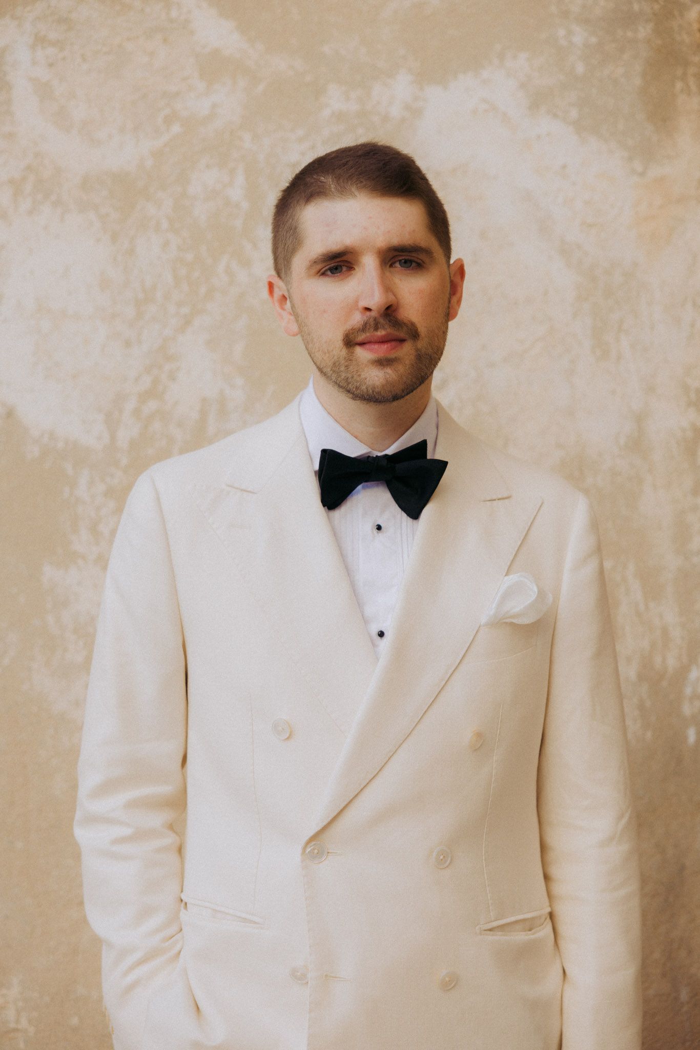 Person wearing a white tuxedo jacket and black bow tie against a beige textured wall.