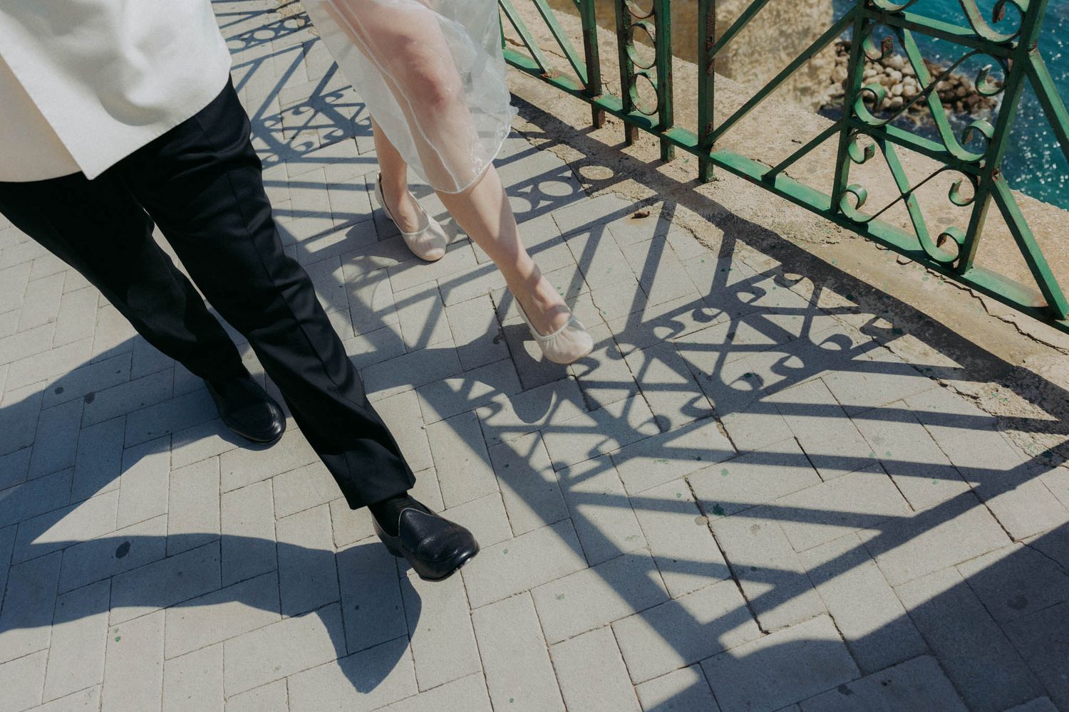 Shadow patterns cast by ornate green railings on a light colored stone balcony floor near the sea.