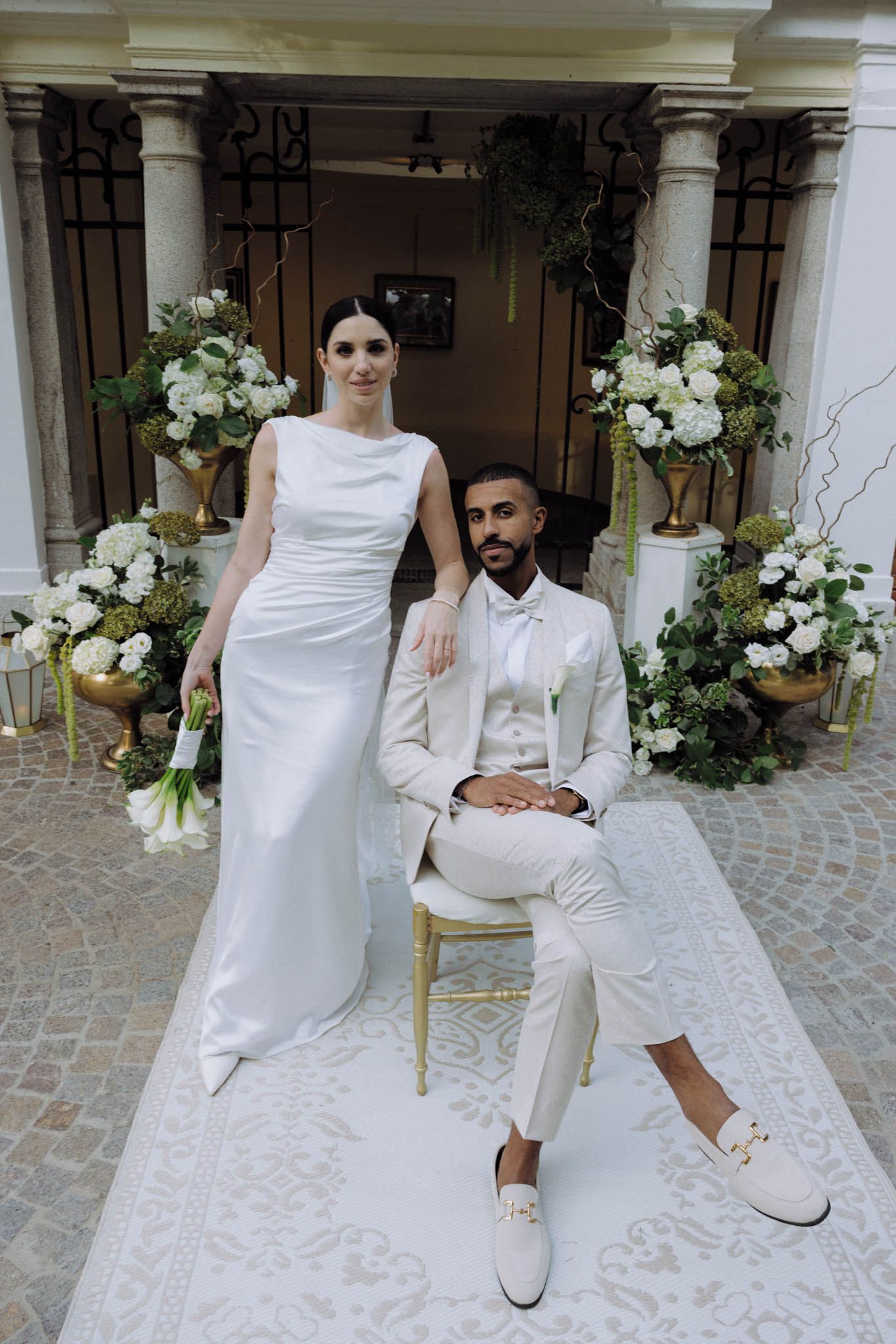A couple in white outfits poses together in an elegant courtyard setting with floral decorations.
