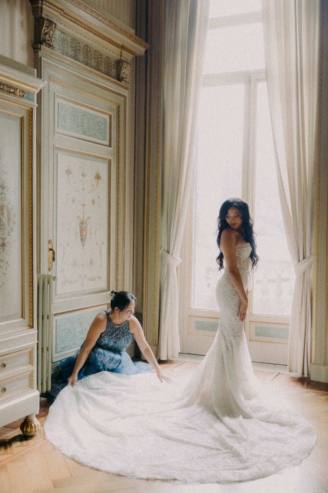 A bride in a flowing white wedding gown with a long train poses elegantly in an ornate room with tall windows.