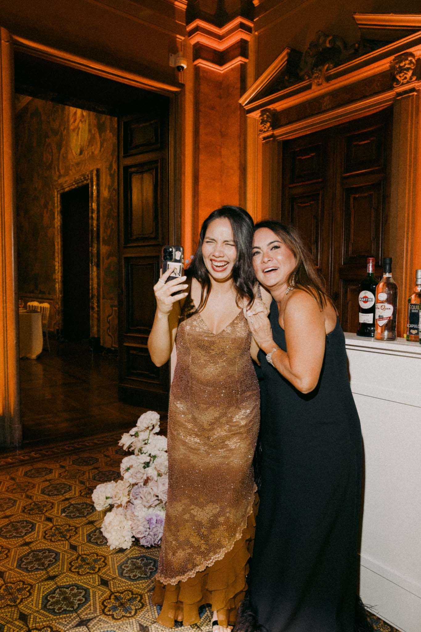 Two people in formal attire pose and laugh together at an indoor event with ornate patterned flooring.